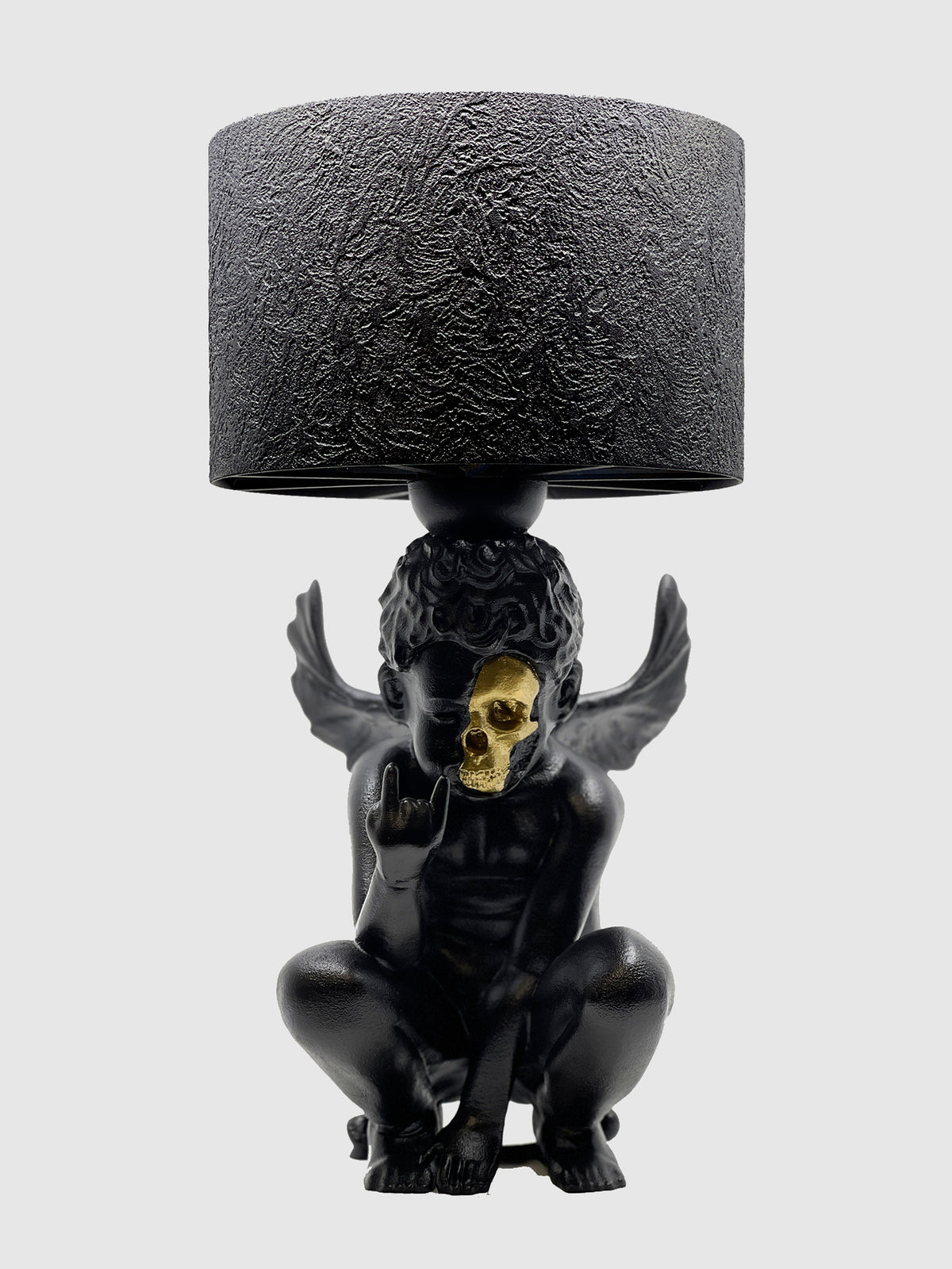 A black lamp with a unique cherub sculpture base featuring a gold-plated skull instead of an eye, paired with a black cylinder lampshade.