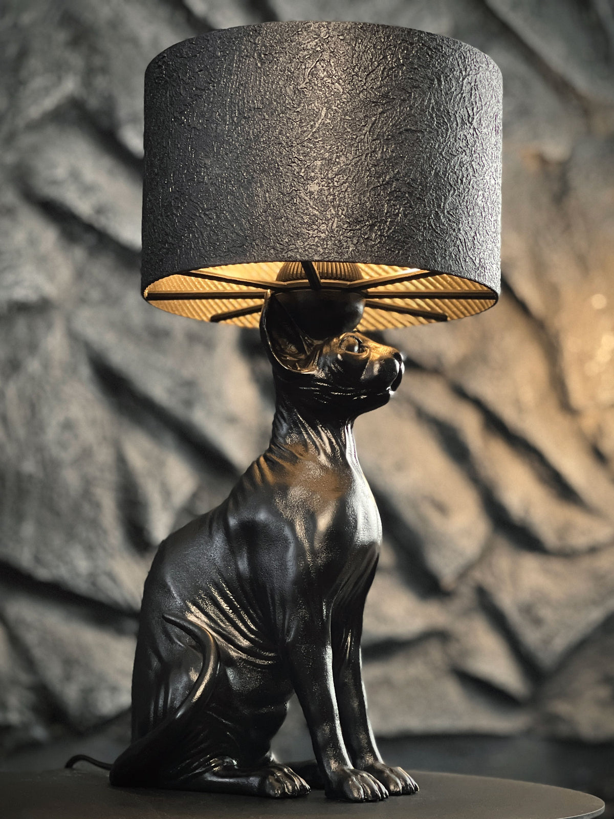 A decorative table lamp featuring a sleek, matte black base sculpted in the shape of a sitting Sphynx cat. The cat’s muscular frame and characteristic skin wrinkles are highlighted by a warm, golden light glowing from beneath a cylindrical, textured black lampshade. The lamp is set against a blurred background of a grey, rocky stone wall.