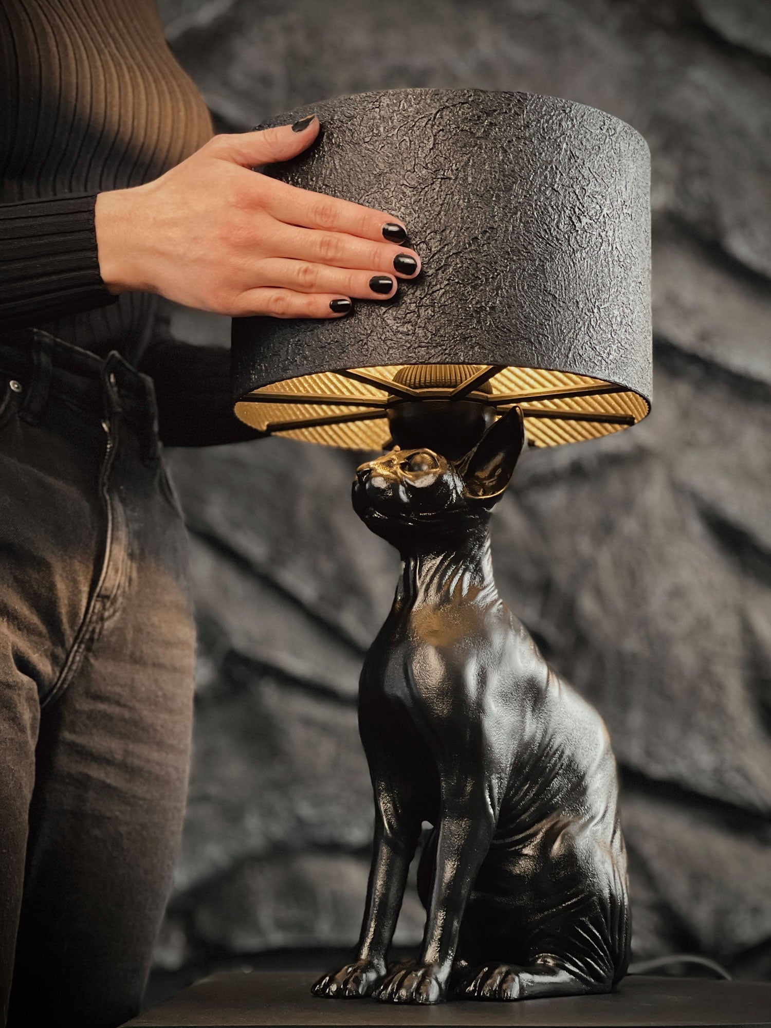 A person with black nail polish adjusts the textured black shade of a matte black Sphynx cat table lamp. The lamp, sculpted with realistic feline details and wrinkles, glows with a warm golden light against a dark, rocky stone wall background.