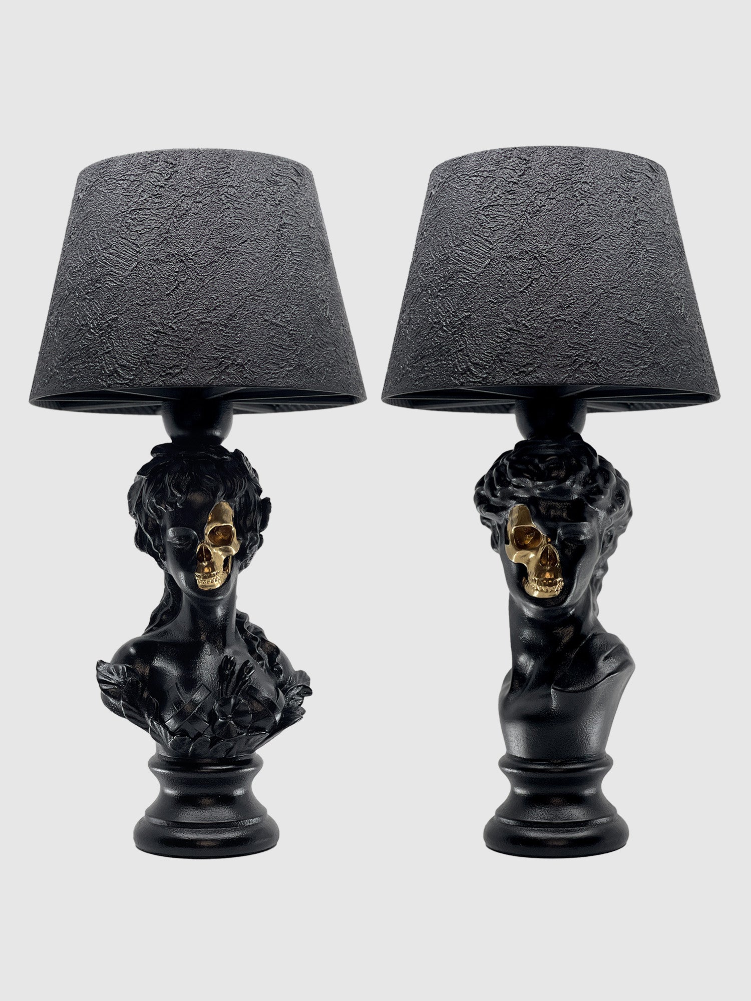 A pair of matte black table lamps featuring busts of Venus and David as bases. Each bust has a section of its face "eroded" or cut away to reveal a shiny, metallic gold skull underneath. Both lamps are topped with matching textured black drum shades against a light gray background.