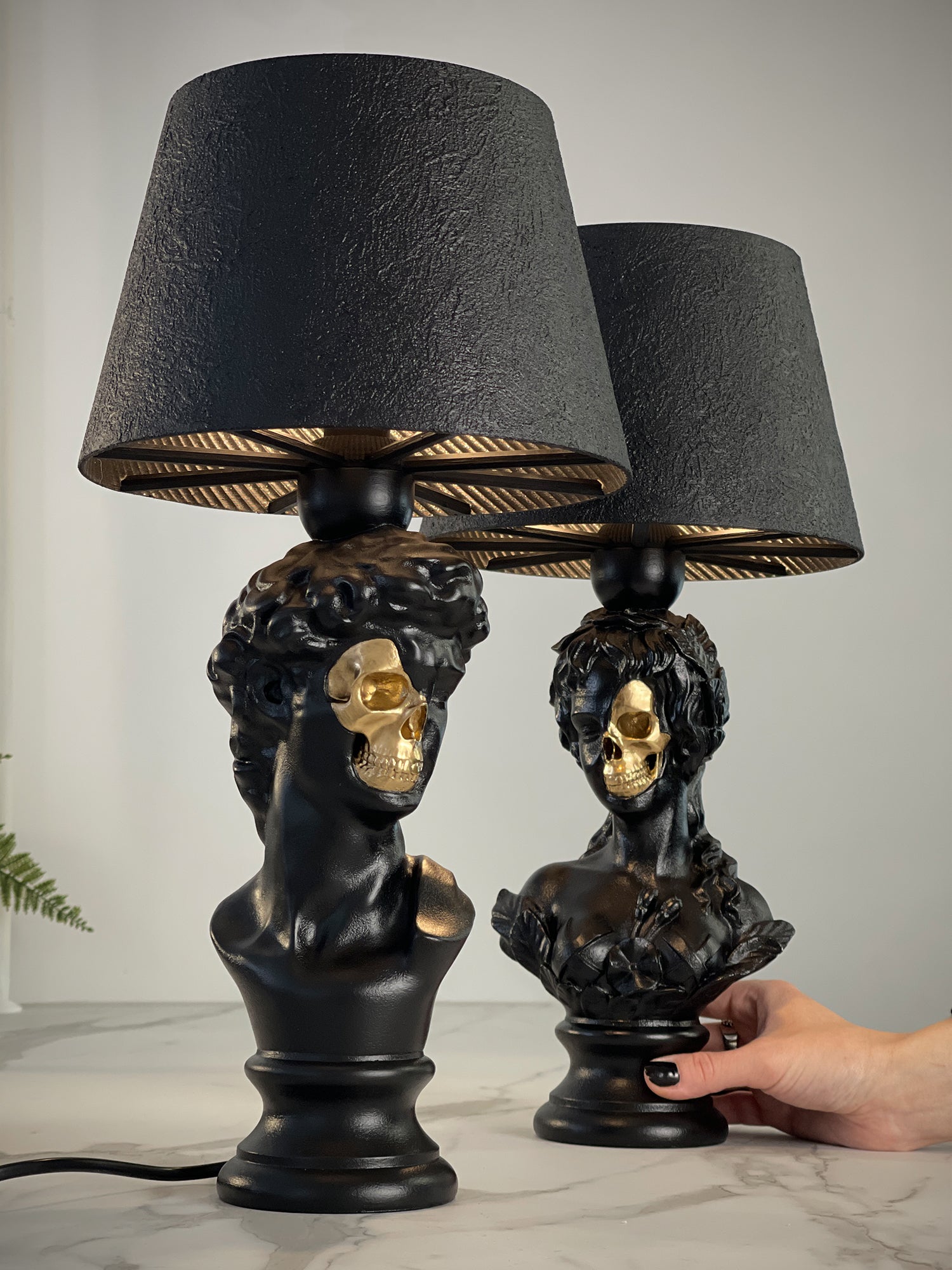 A pair of "Venus & David" Renaissance bust lamps in a matte black finish. Each classical bust features a dramatic anatomical detail: a metallic gold skull revealed on one side of the face. The lamps are topped with textured black tapered shades with gold interior ribbing, shown on a light marble surface.