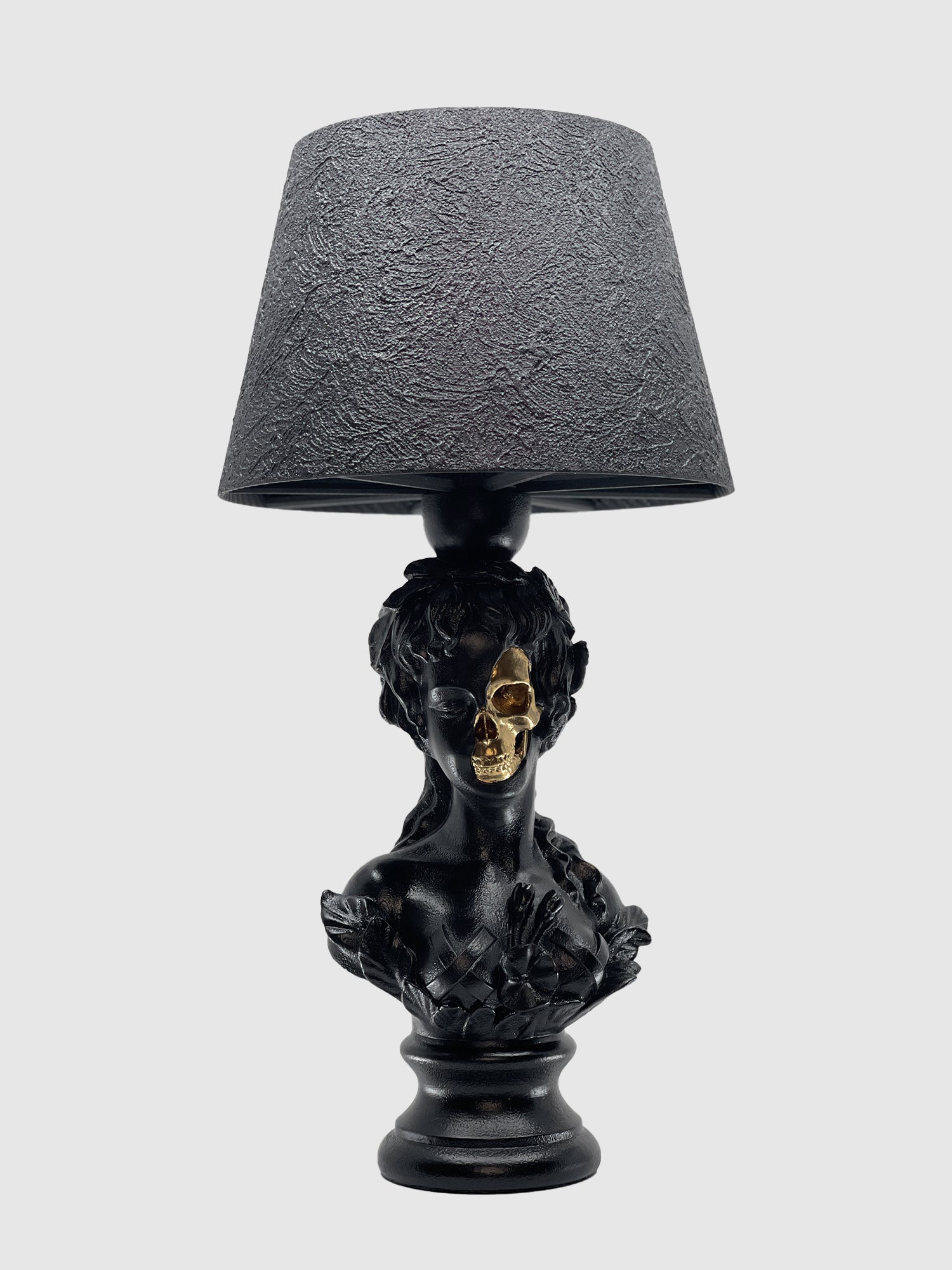 A front view of the matte black table lamp featuring a bust of Venus. A portion of her face is removed to reveal a metallic gold skull beneath. The lamp is topped with a matching textured black drum shade and set against a light gray background.