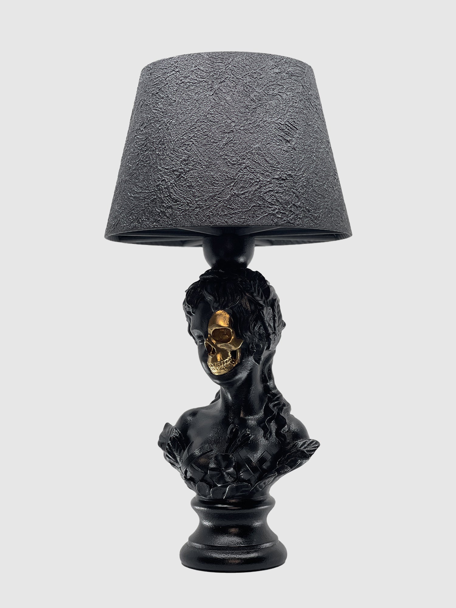 A matte black table lamp featuring a bust of Venus as the base. A portion of her face is removed to reveal a metallic gold skull beneath. The lamp is topped with a matching textured black drum shade and set against a light gray background.