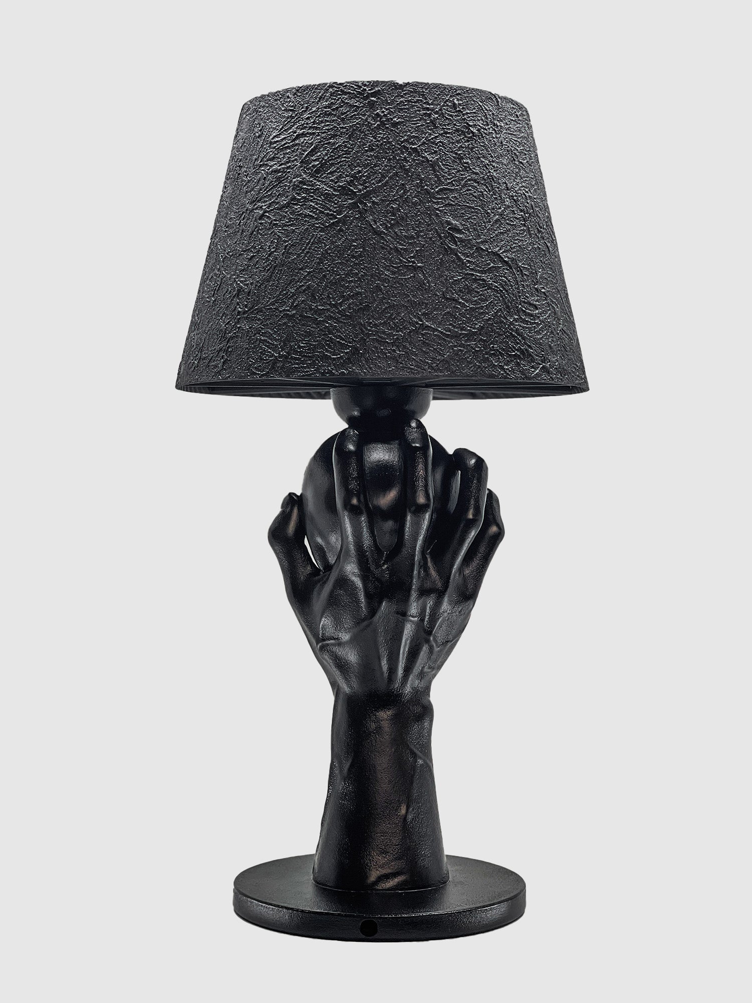 A back view of the "To Be or Not To Be" matte black table lamp by Horror Romance Design. The sculptural base shows the detailed back of a hand and wrist gripping a skull, topped with a textured black shade against a white background.