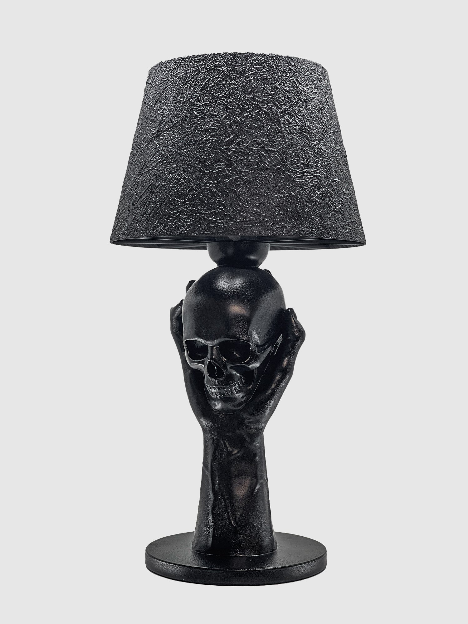 A matte black table lamp featuring a sculptural base of two hands holding a human skull, titled "To Be or Not To Be" by Horror Romance Design. The lamp is topped with a textured, black fabric shade and is displayed on a clean white background.