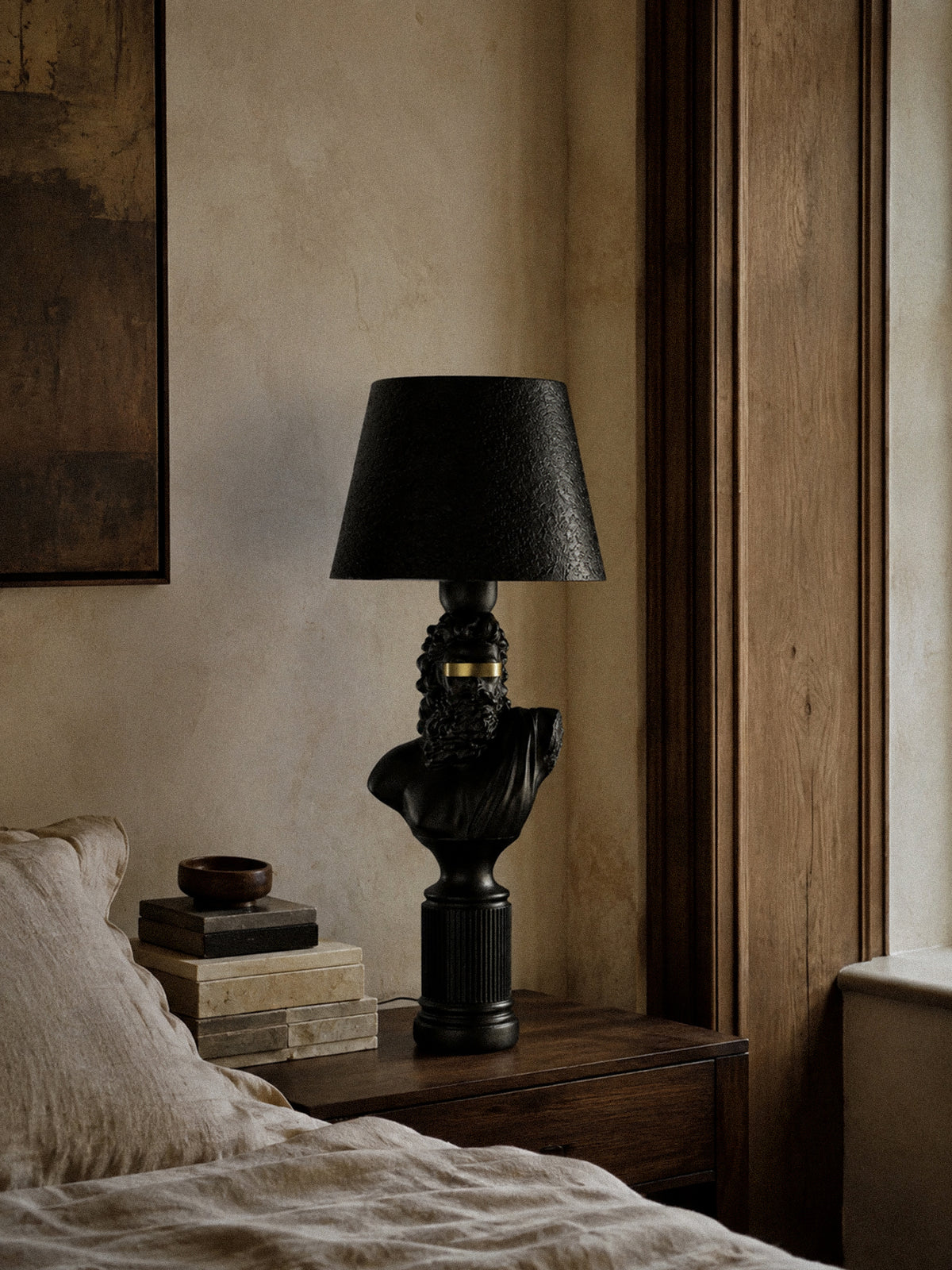 Blind Zeus black and gold bust lamp on a wooden bedside cabinet beside a window and textured walls.