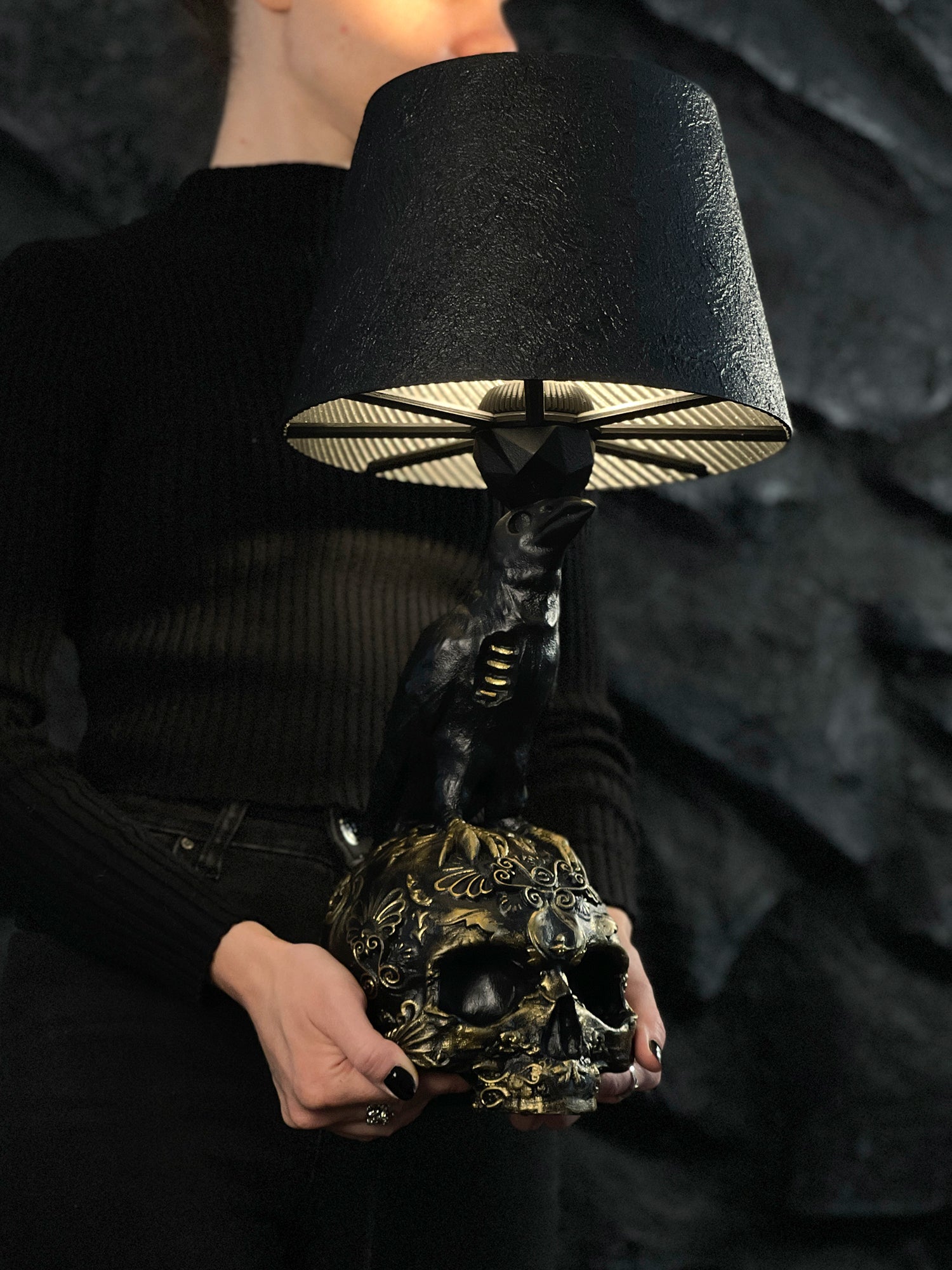 A person dressed in black holds the "Raven Skull – Gold Baroque Filigree Desk Lamp" against a dark, craggy stone wall. The lamp features a large skull base adorned with intricate gold baroque patterns, topped with a black raven. A textured black shade completes the piece, glowing with light that highlights its golden-ribbed interior.
