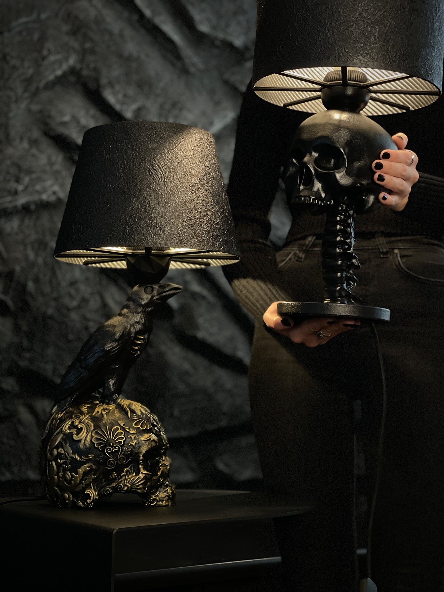 A side-by-side display of two gothic desk lamps against a dark stone wall. On the left, the "Raven Skull – Gold Baroque Filigree Desk Lamp" features a black raven perched atop a skull adorned with intricate gold filigree. On the right, a person holds the "Skull Spine – Anatomical Desk Lamp," which consists of a matte black skull resting on a detailed spinal column base. Both lamps are finished with matching textured black shades that reveal a golden, ribbed interior.