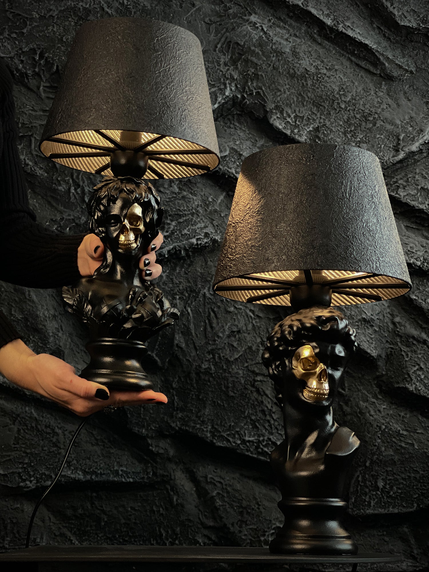 A product shot of the "Venus & David Set – Half-Skull Renaissance Bust Lamps" against a craggy, dark grey stone wall. The matte black Venus and David busts, each featuring a polished gold half-skull face, are illuminated from beneath their textured black lampshades. A person's hands with black nail polish are shown holding the Venus lamp, emphasizing the set's scale and gothic aesthetic.