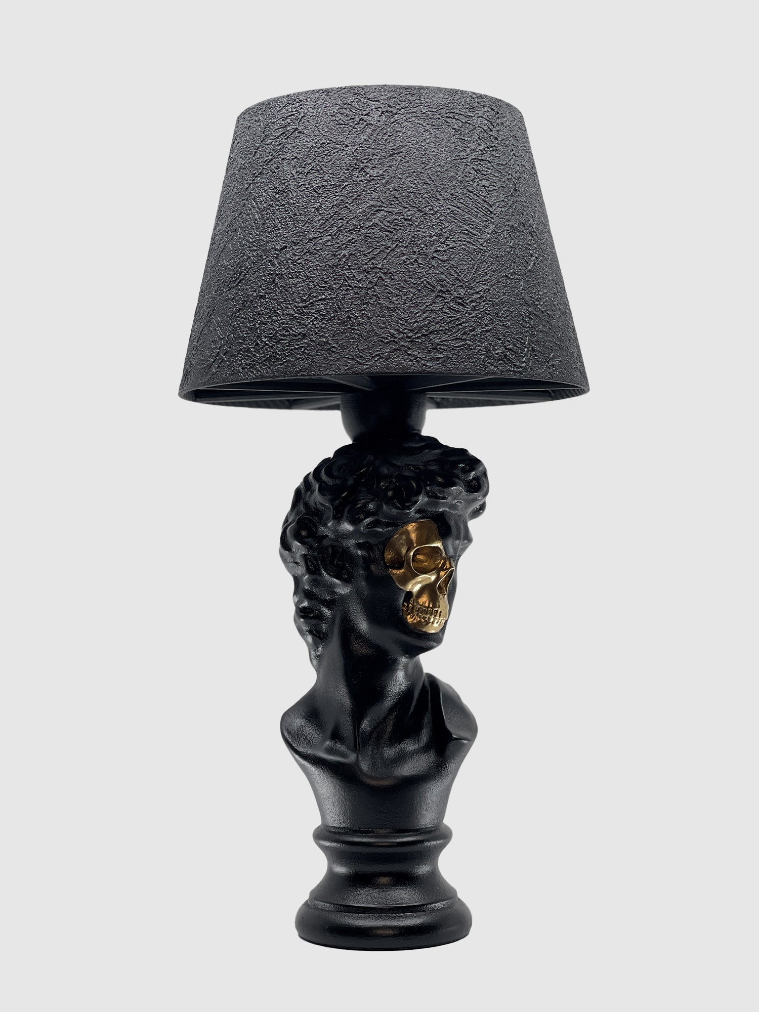 A three-quarter view of the matte black table lamp featuring a bust of David. A section of the face is cut away to reveal a shiny, metallic gold skull underneath. The lamp is topped with a textured black drum shade against a light gray background.