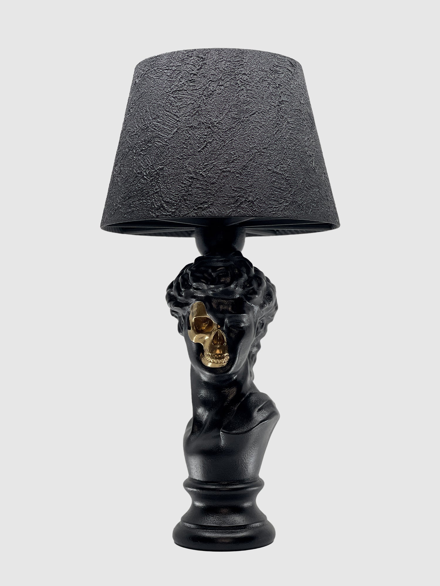 A front view of the matte black table lamp featuring a bust of David. A section of the face is cut away to reveal a gleaming gold skull underneath. The lamp is topped with a matching textured black drum shade and set against a light gray background.
