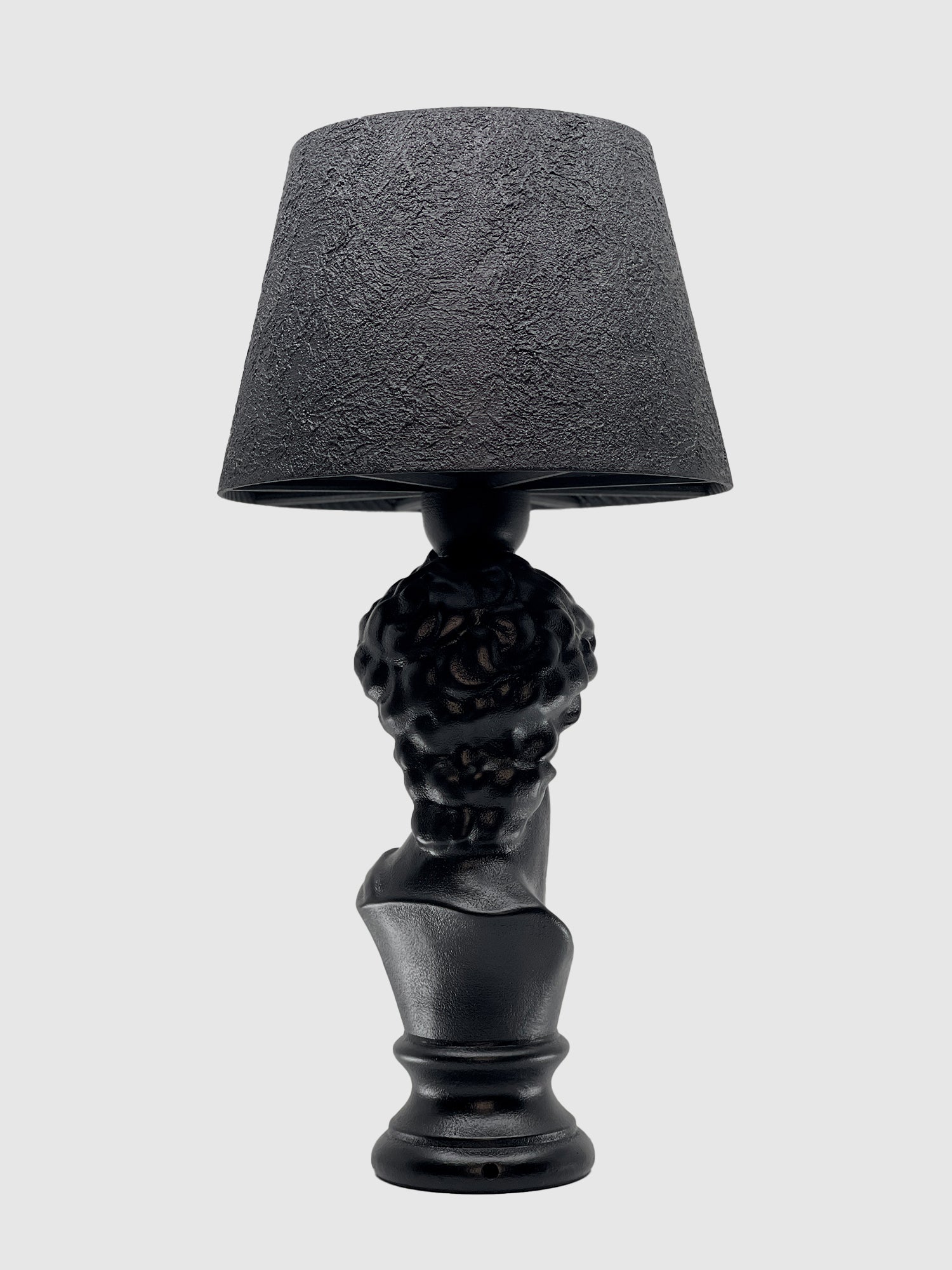 A back view of the matte black table lamp featuring the bust of David. The base shows the detailed, curly hair of the sculpture, and the lamp is topped with a matching textured black drum shade against a light gray background.