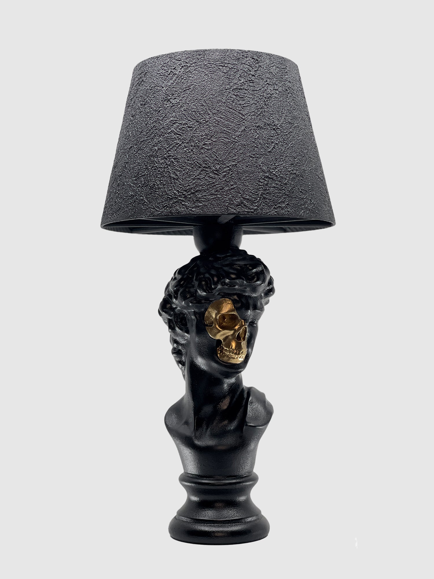 A matte black table lamp featuring a bust of Michelangelo's David as the base. A section of the face is carved away to reveal a gleaming gold skull underneath. The lamp is topped with a black textured drum shade and set against a solid light gray background.