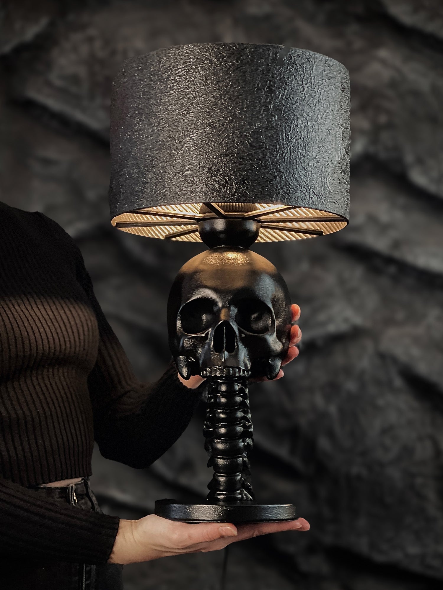 A person holding a matte black table lamp featuring a base shaped like a human skull resting on a spinal column. The lamp has a textured dark grey drum shade and is set against a dark, craggy stone background.