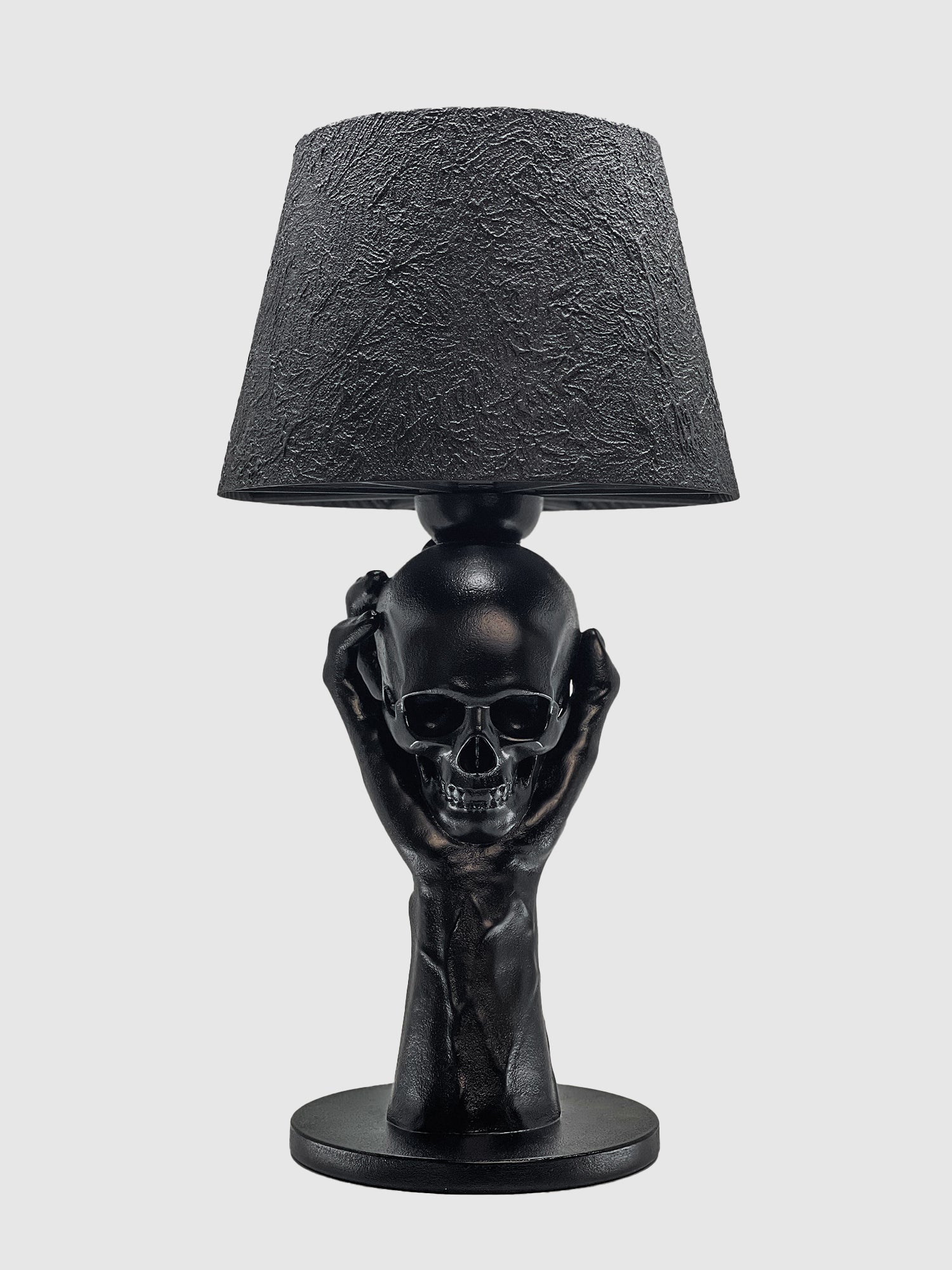 A front-facing view of the "To Be or Not To Be" matte black table lamp by Horror Romance Design. The sculptural base consists of a hand holding a human skull, supporting a conical lampshade with a heavily textured, cracked-earth finish.