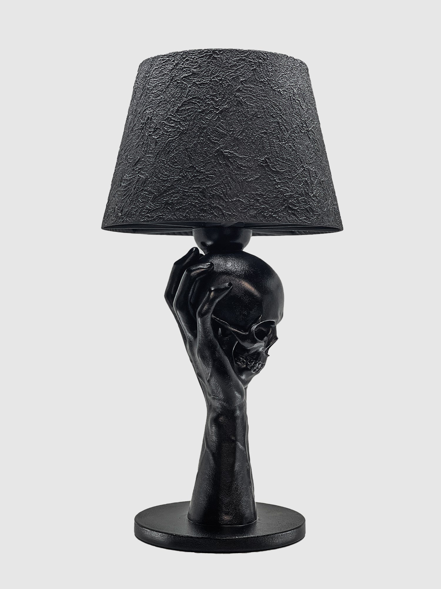 A three-quarter view of the matte black "To Be or Not To Be" table lamp by Horror Romance Design. The sculptural base depicts a hand firmly gripping a human skull from the side and back, supporting a conical lamp shade with a heavy, cracked texture against a plain white background.