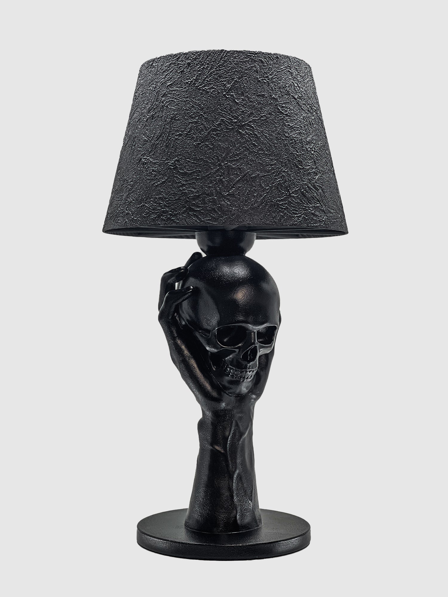 A matte black table lamp featuring a sculptural base of a hand holding a human skull. The lamp is topped with a textured, cracked-finish black shade and set against a plain light gray background.