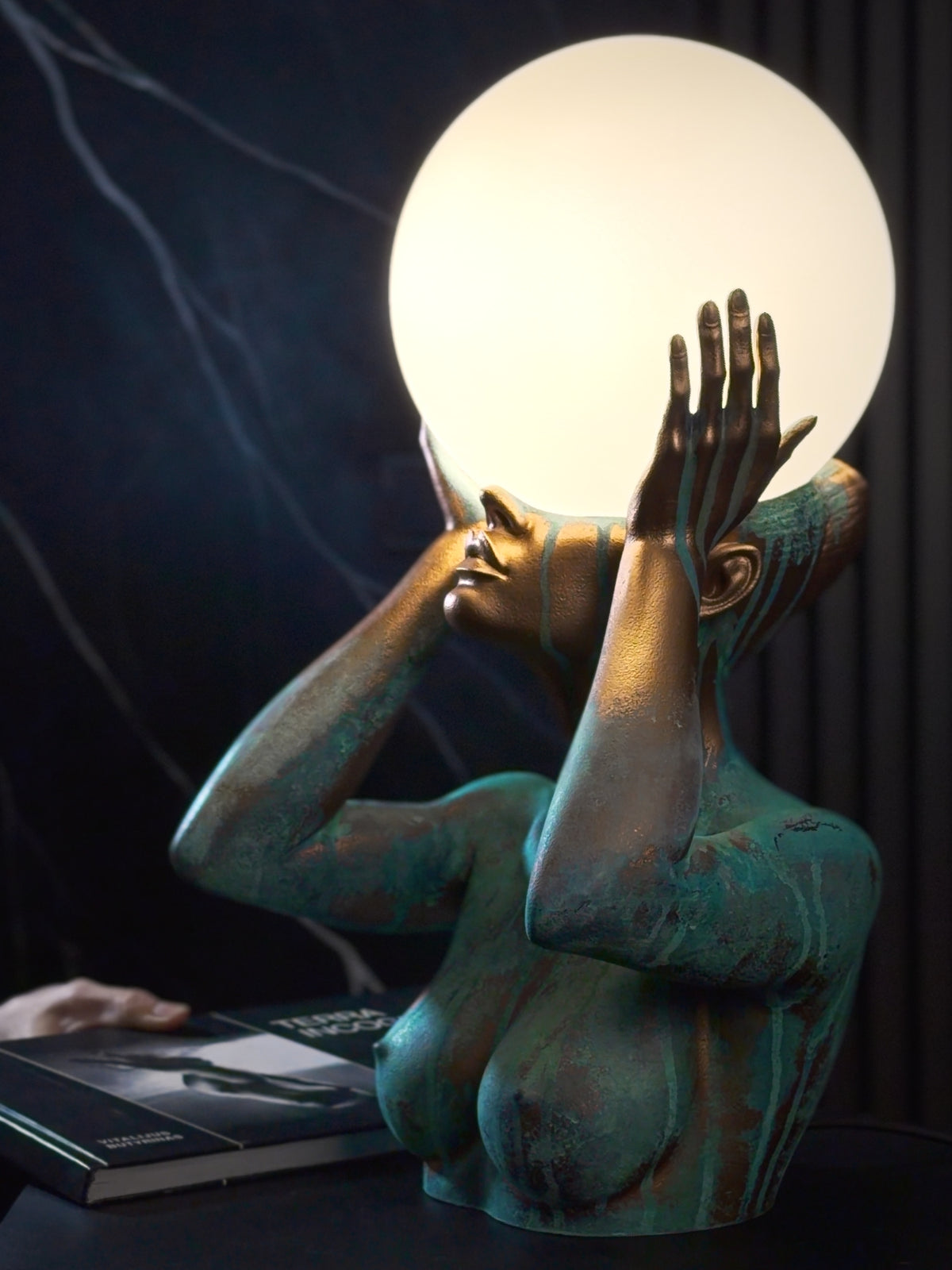 Video of SAULĖ ORACLE sculptural lamp, featuring a female figure holding a glowing globe between raised hands, with close-up and side views of its copper finish and turquoise patina.