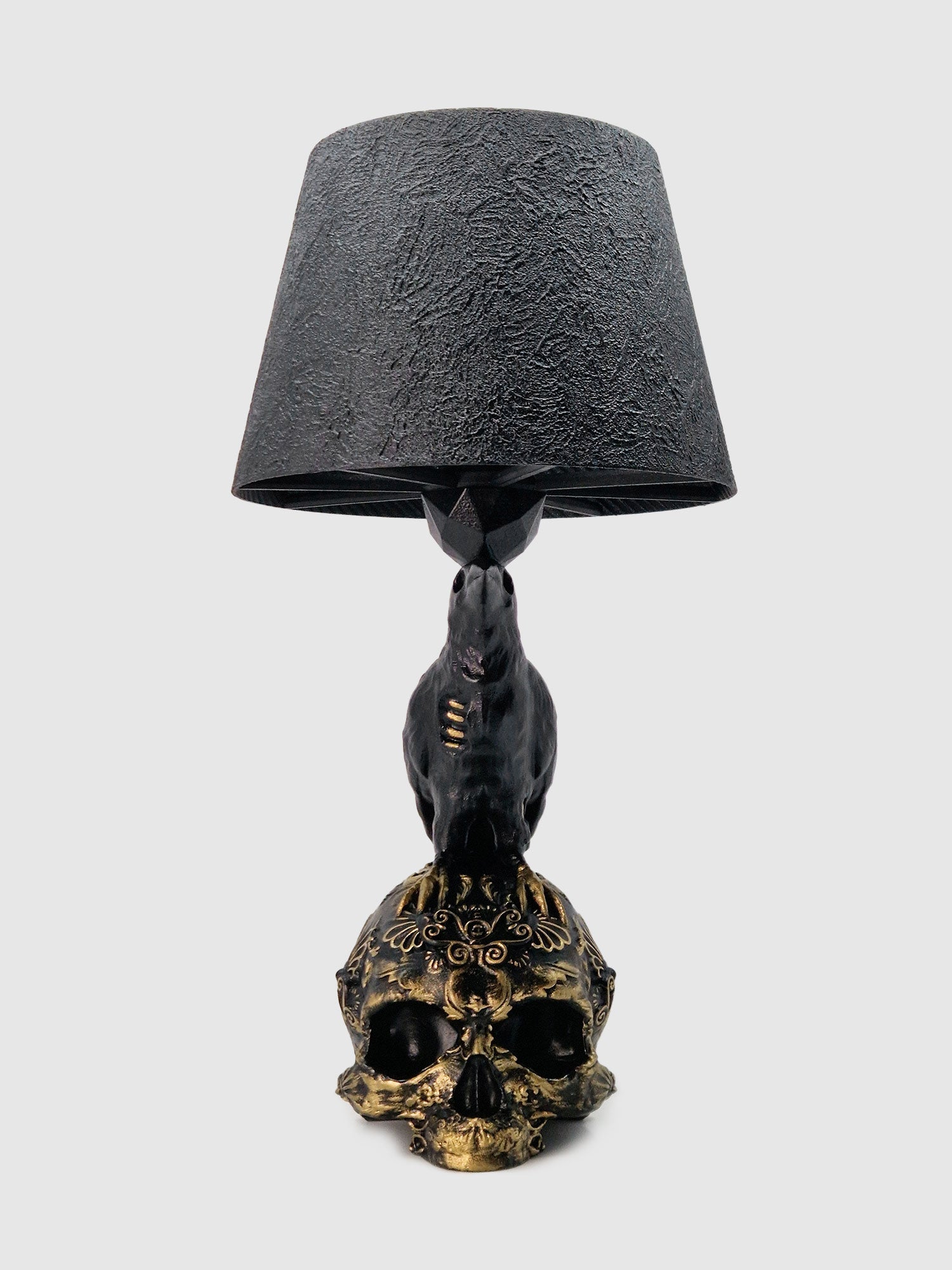 Front view of the Raven Skull lamp: a black raven perched on a gold baroque filigree skull base designed by Horror Romance.