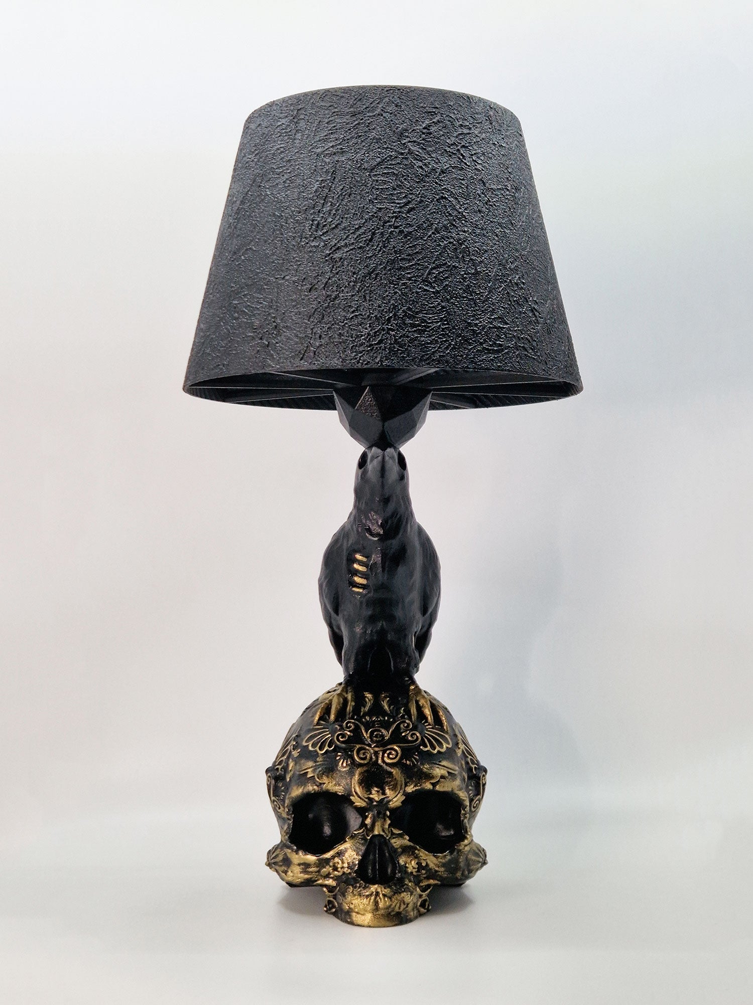 Front view of the Raven Skull lamp: a black raven perched on a gold baroque filigree skull base.