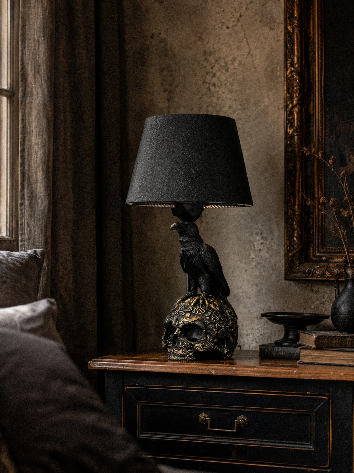 Raven Skull black and gold table lamp on a dark bedside cabinet beside an aged wall and framed artwork.
