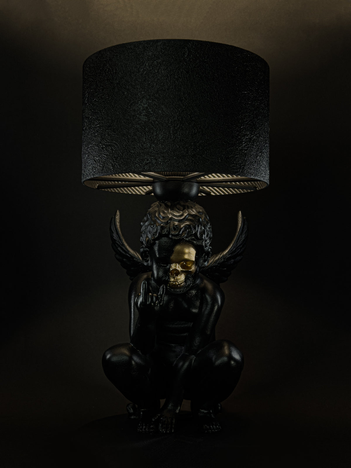 Front view of the illuminated Rebel Angel lamp with a black cherub, gold half-skull face and raised wings.