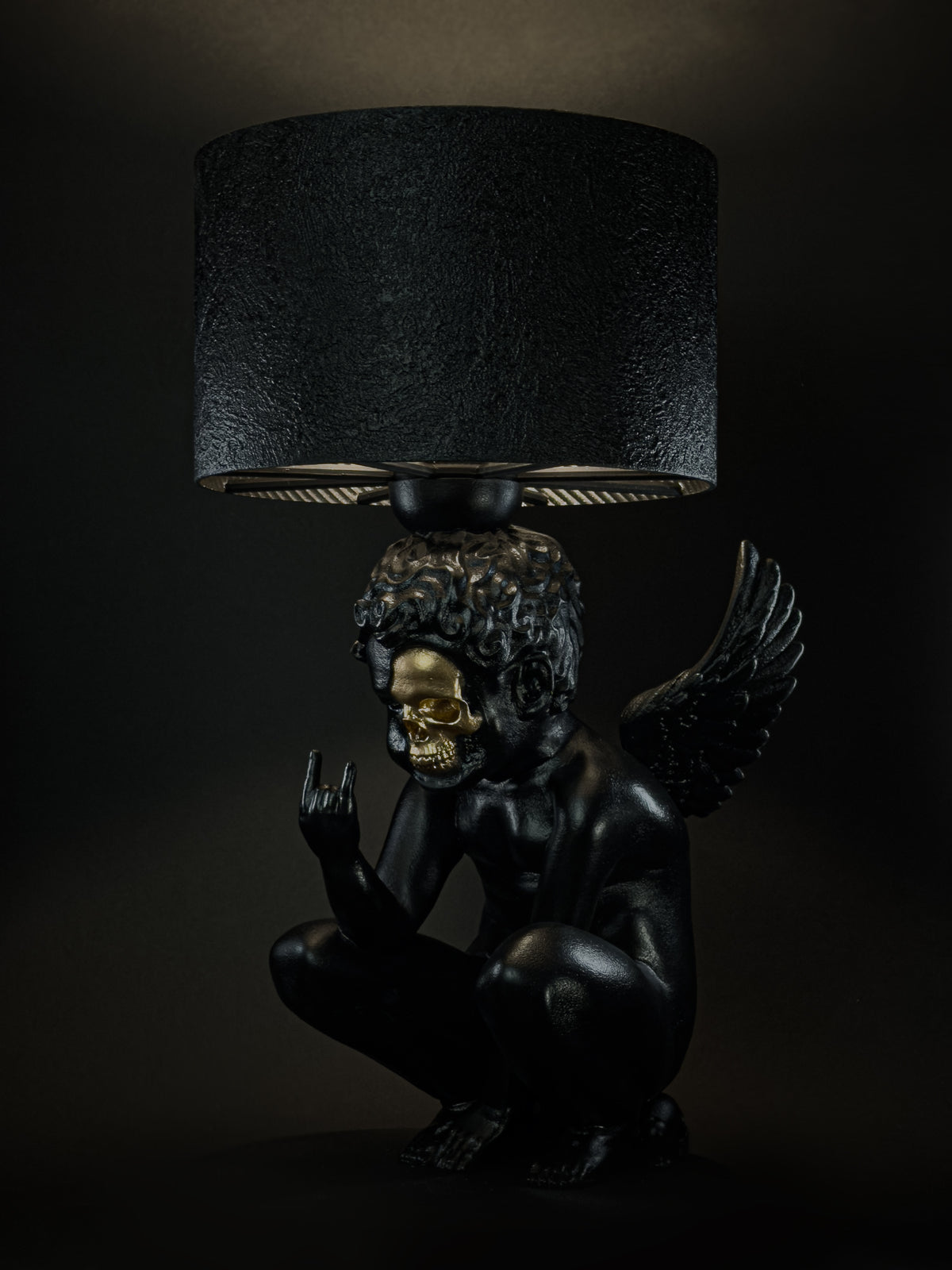 Side view of the illuminated Rebel Angel lamp with a black cherub, gold skull detail and raised hand.