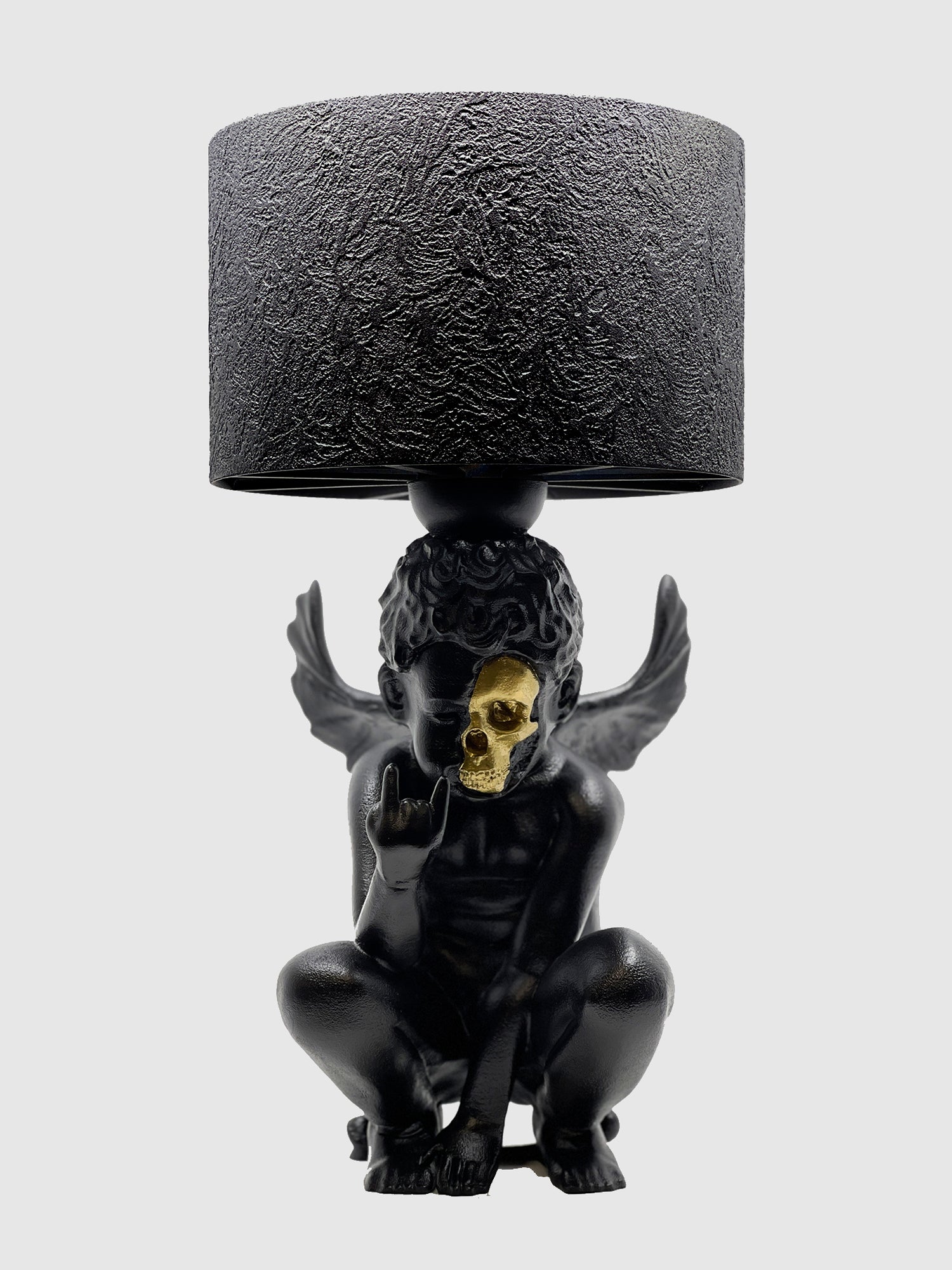 Front view of the Rebel Angel lamp: matte black crouching cherub with a gold-plated skull face and rock on hand gesture.