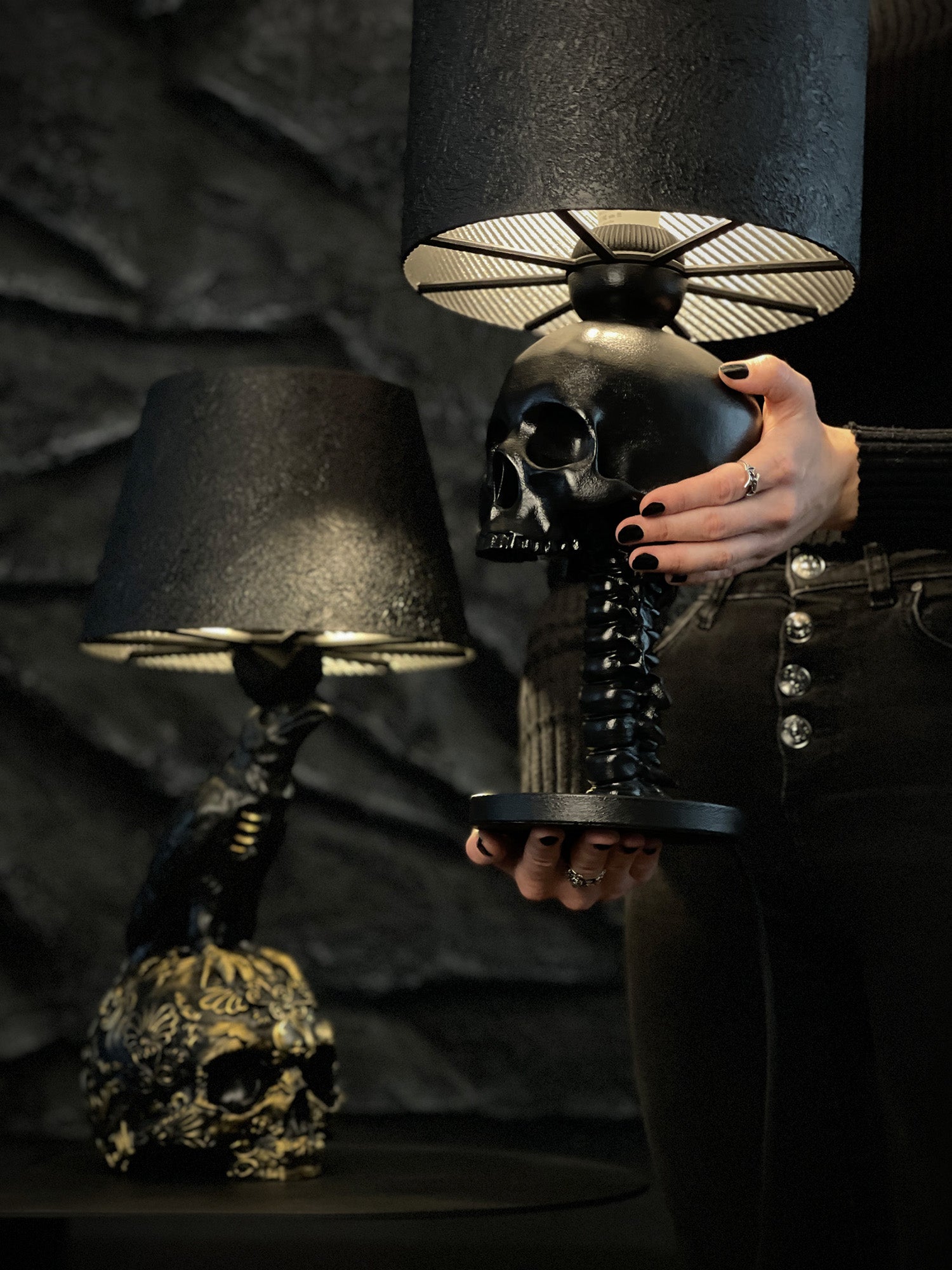 A moody close-up shot of two gothic desk lamps against a dark, craggy stone wall. In the foreground, a person with black nail polish holds the "Skull Spine – Anatomical Desk Lamp," featuring a matte black skull mounted on a detailed spinal column. To the left, the "Raven Skull – Gold Baroque Filigree Desk Lamp" is visible, showing a black raven perched on a skull with ornate gold filigree. Both lamps feature textured black shades with glowing, golden-ribbed interiors.
