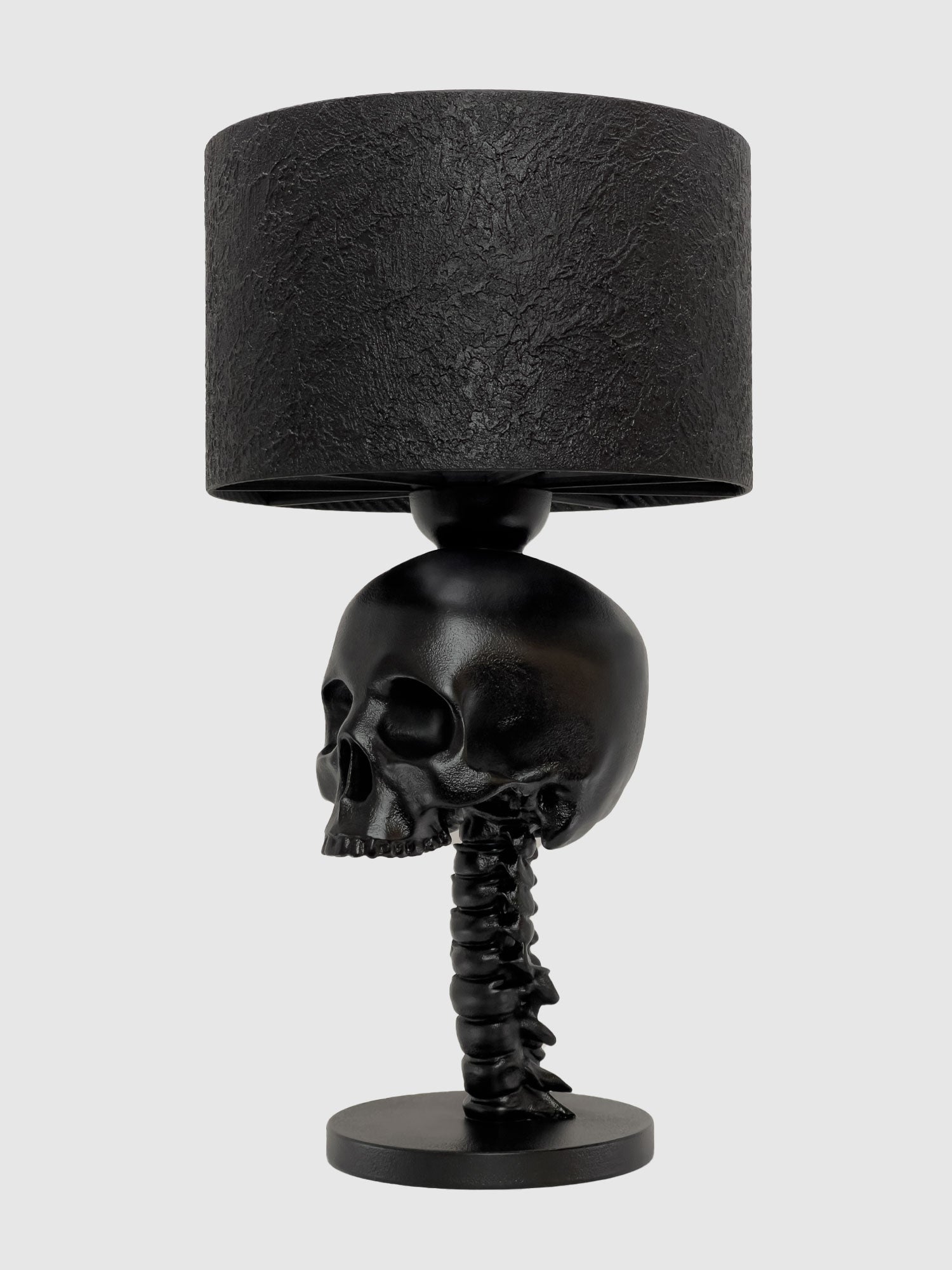 A side profile view of the "Skull Spine" anatomical desk lamp in a satin black finish. The realistic human skull faces left, perched atop a detailed cervical spinal column that serves as the lamp's base, topped with a textured black drum shade, created by Horror Romance.
