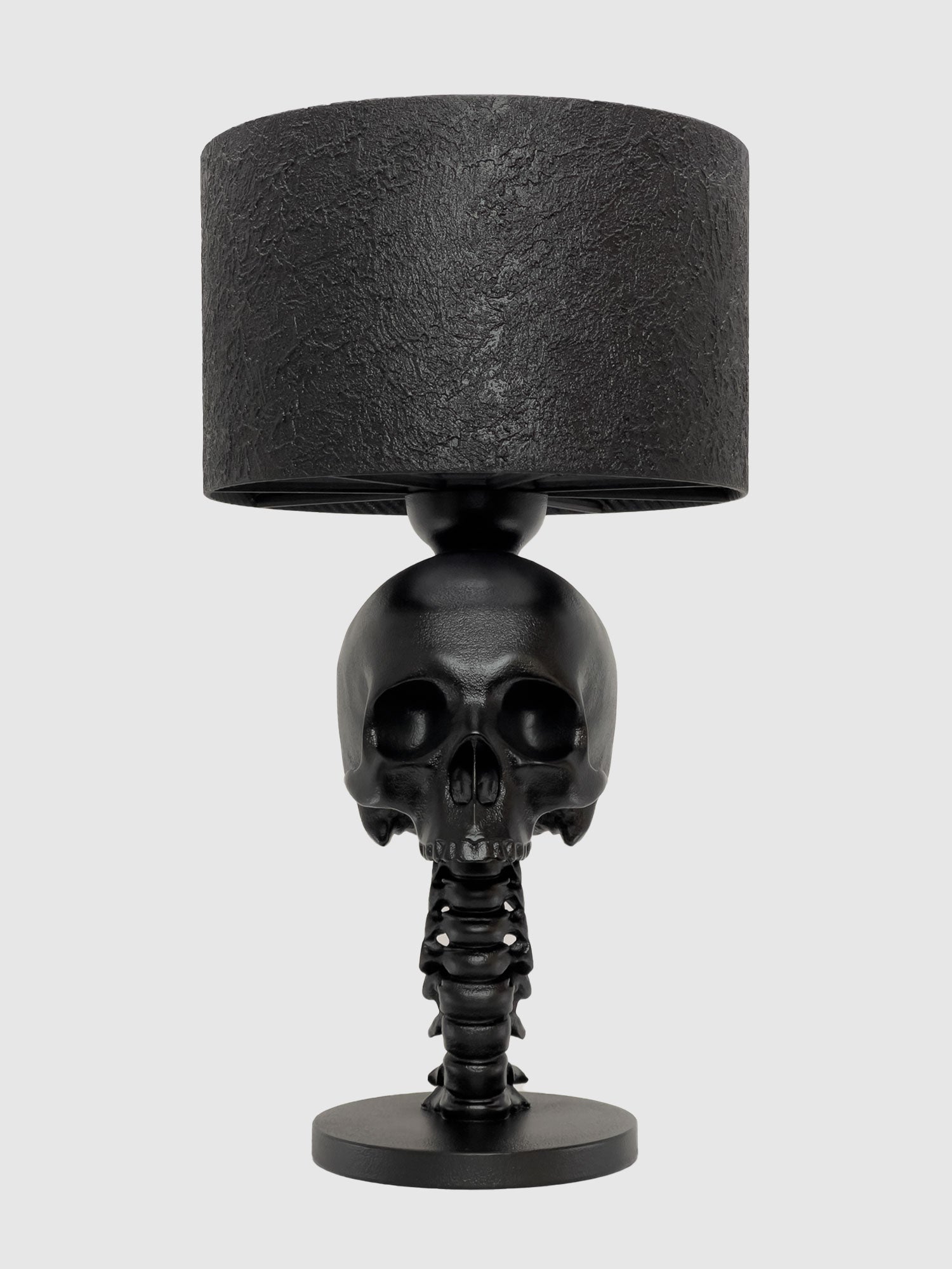 A direct front-facing view of the matte black Skull Spine Lamp. The medical-grade skull sculpture stares forward, supported by a meticulously detailed vertebrae column and a circular base, showcasing a monochromatic gothic aesthetic, designed by Horror Romance.