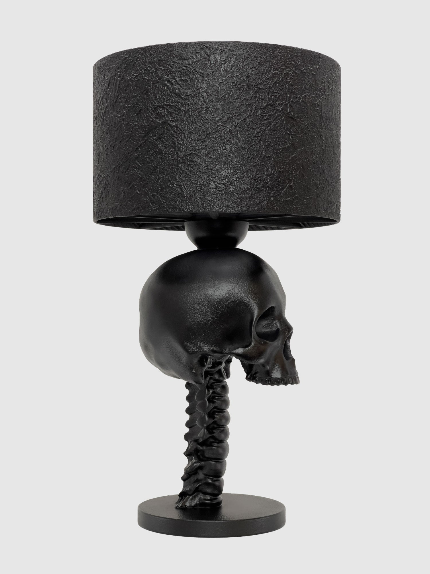 A right-side profile view of the Skull Spine Anatomical Desk Lamp. The skull faces right, emphasizing the organic micro-texture of the concrete-coated finish and the structural beauty of the vertebral base, design by Horror Romance.