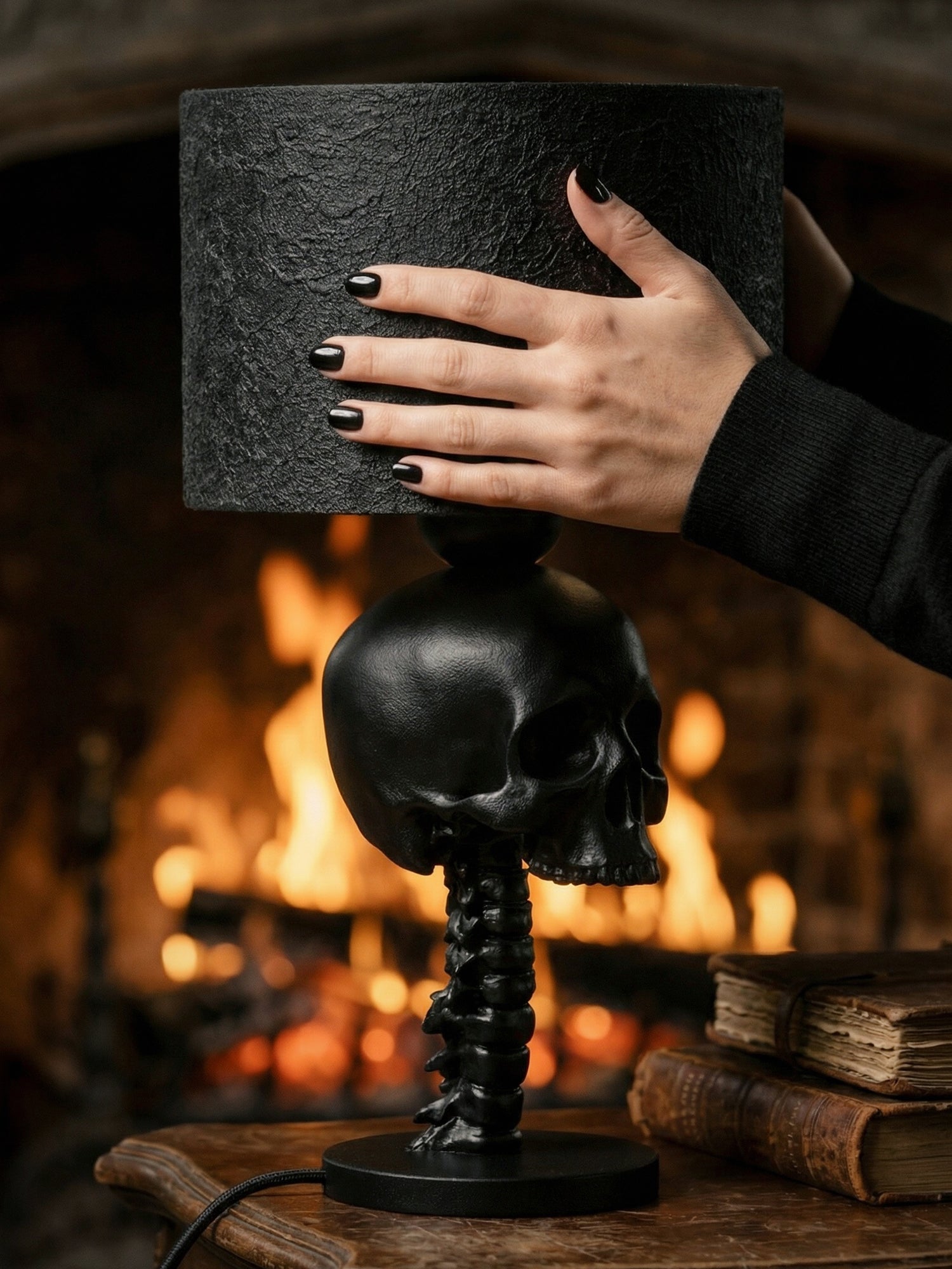 A matte black gothic-style table lamp with a base shaped like an anatomical spinal column and a human skull. A person’s hand with black nail polish rests on the textured, cylindrical black lampshade. In the background, a warm fire glows in a fireplace, and a couple of weathered, leather-bound books sit on the wooden table beside the lamp.