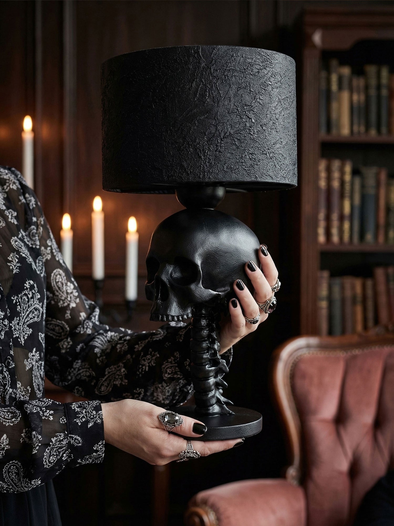 A person with black nail polish and ornate silver rings holds a matte black skull table lamp with a textured drum shade in a dimly lit, gothic-style library.