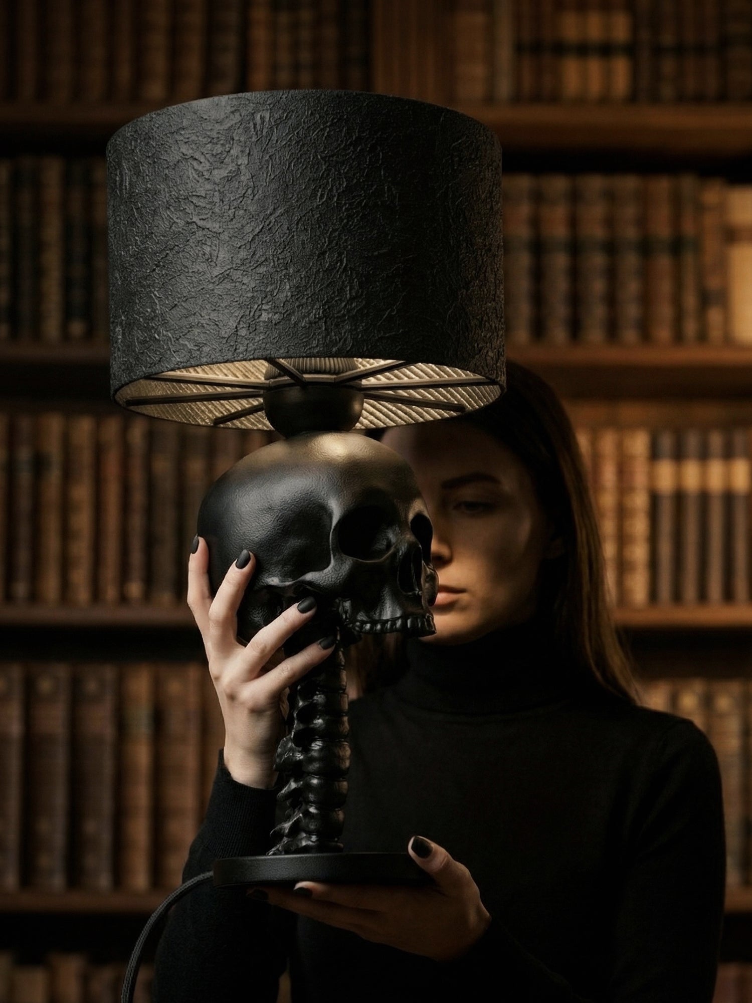 A person wearing a black turtleneck holds the matte black skull and spine lamp with both hands. The lamp is positioned in front of them, partially obscuring their face. The background is a dark library filled with floor-to-ceiling bookshelves, creating a "dark academia" aesthetic.