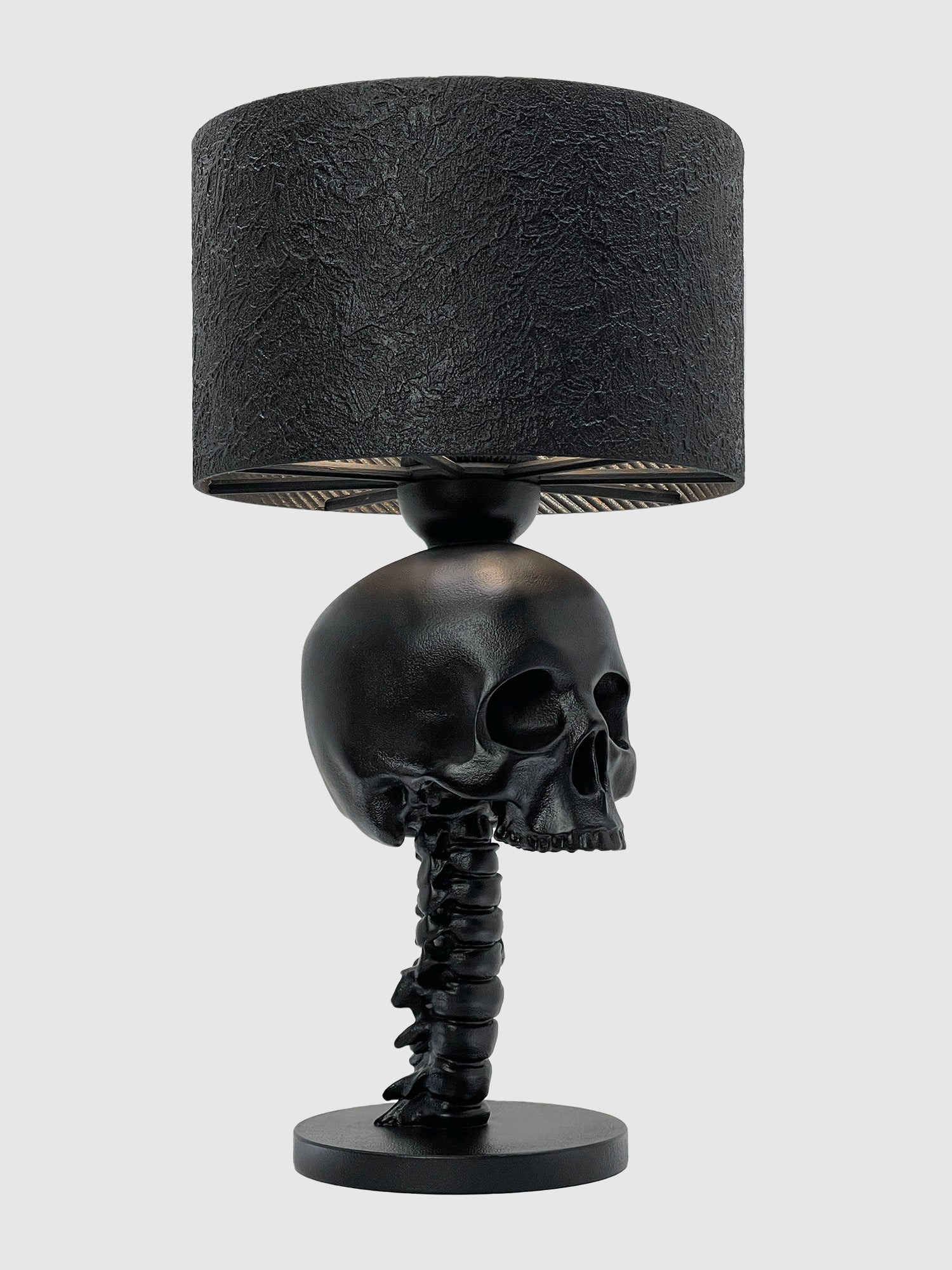 A professional product shot of a gothic table lamp against a solid light gray background. The lamp features a matte black finish with a base sculpted into a detailed spinal column that supports a human skull. It is topped with a black lampshade that has a heavily textured, bark-like surface.