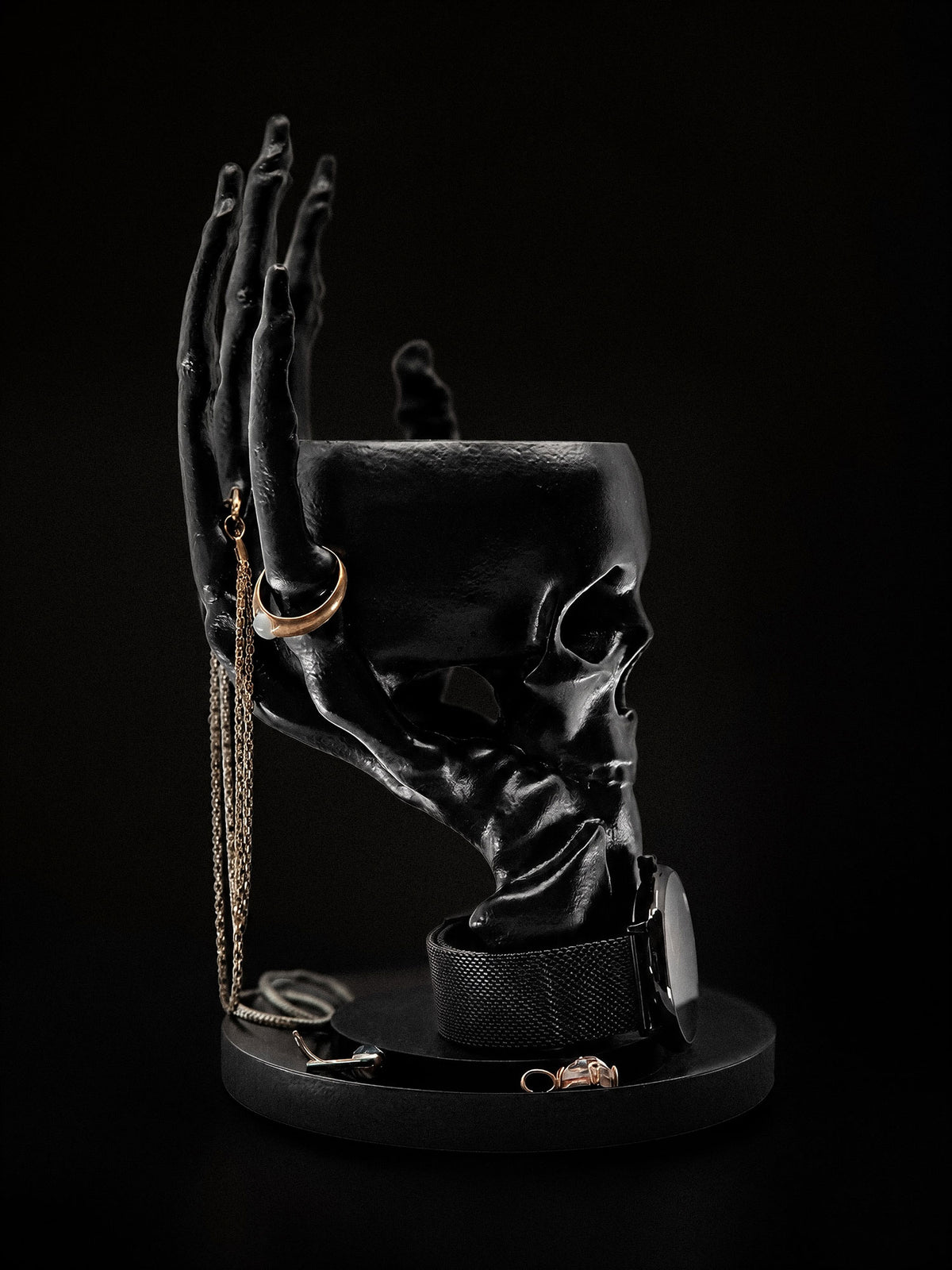 Side view of The Offering skull jewelry bowl supported by a black skeletal hand, with rings on the fingers.