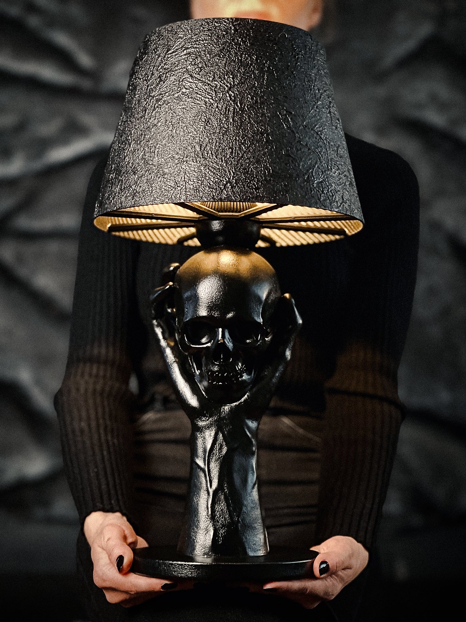 A gothic-style table lamp called "To Be or Not To Be" featuring a matte black sculpted base of two hands holding a human skull, topped with a textured black lampshade being held by a person against a dark rocky background.