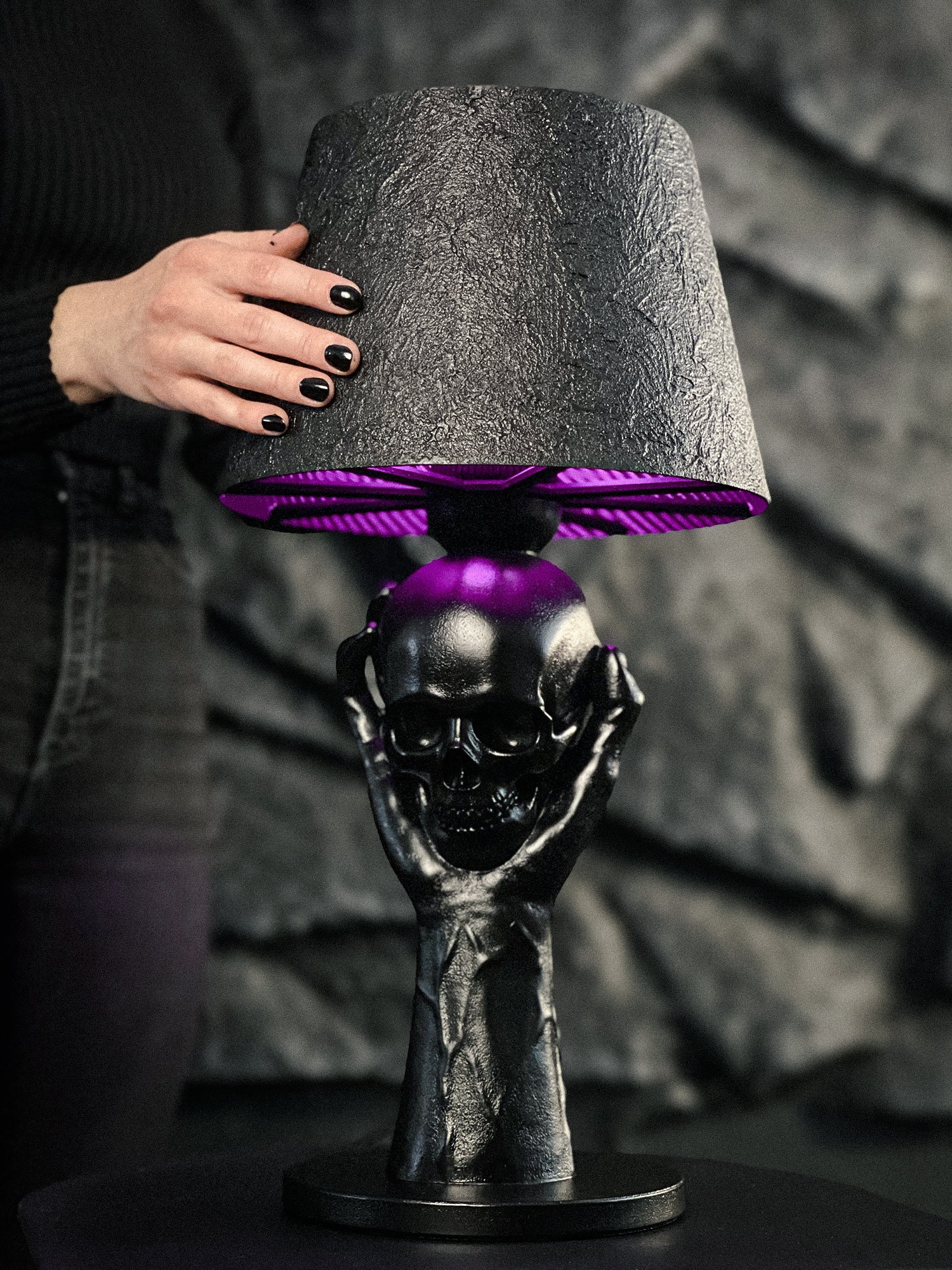 A gothic-inspired To Be or Not To Be – Sculpted Hand & Skull Lamp emitting a vibrant purple glow from beneath a textured black lampshade. The base is a detailed matte black sculpture of hands holding a human skull, shown being held by a person with black nail polish against a dark, craggy backdrop.