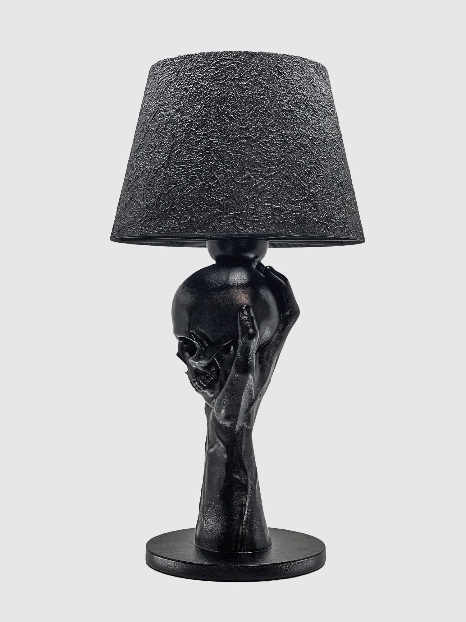 A side profile view of the "To Be or Not To Be" table lamp by Horror Romance Design. The matte black base depicts a hand reaching up to hold a human skull, supporting a textured black lamp shade against a clean white background.