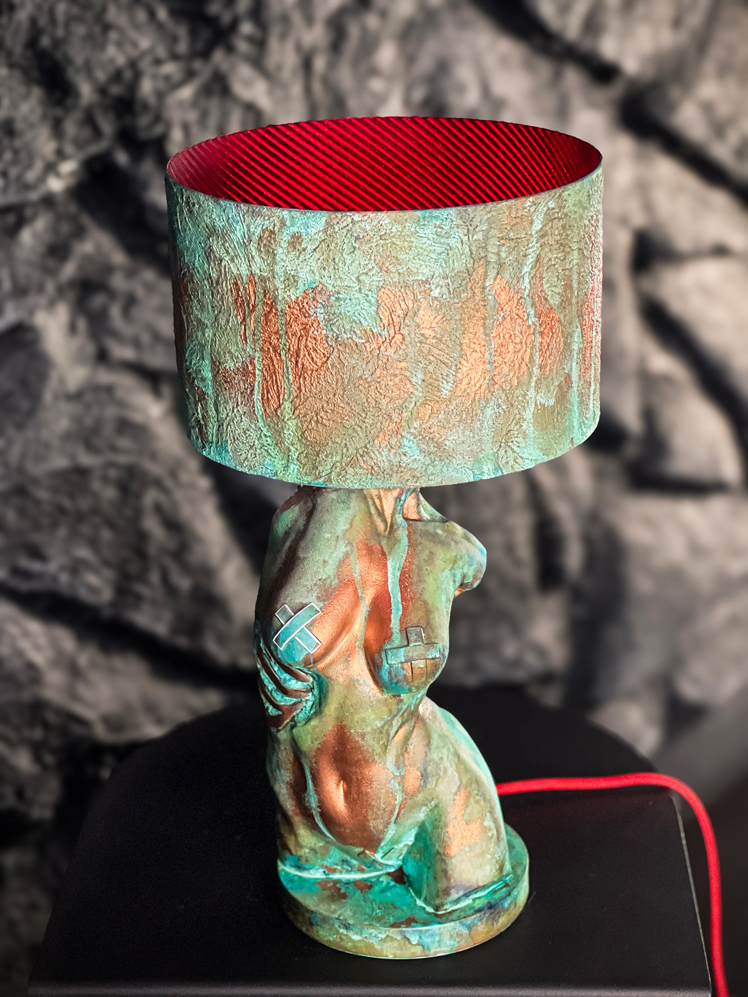 An artistic table lamp with a turquoise verdigris and copper-patinated female torso base. The sculpture features metallic tape accents on the breasts and visible ribs on the side. It is topped with a matching textured drum shade that reveals a vibrant, ribbed red interior. The lamp rests on a black surface against a dark, rocky background, with a bright red power cord visible.