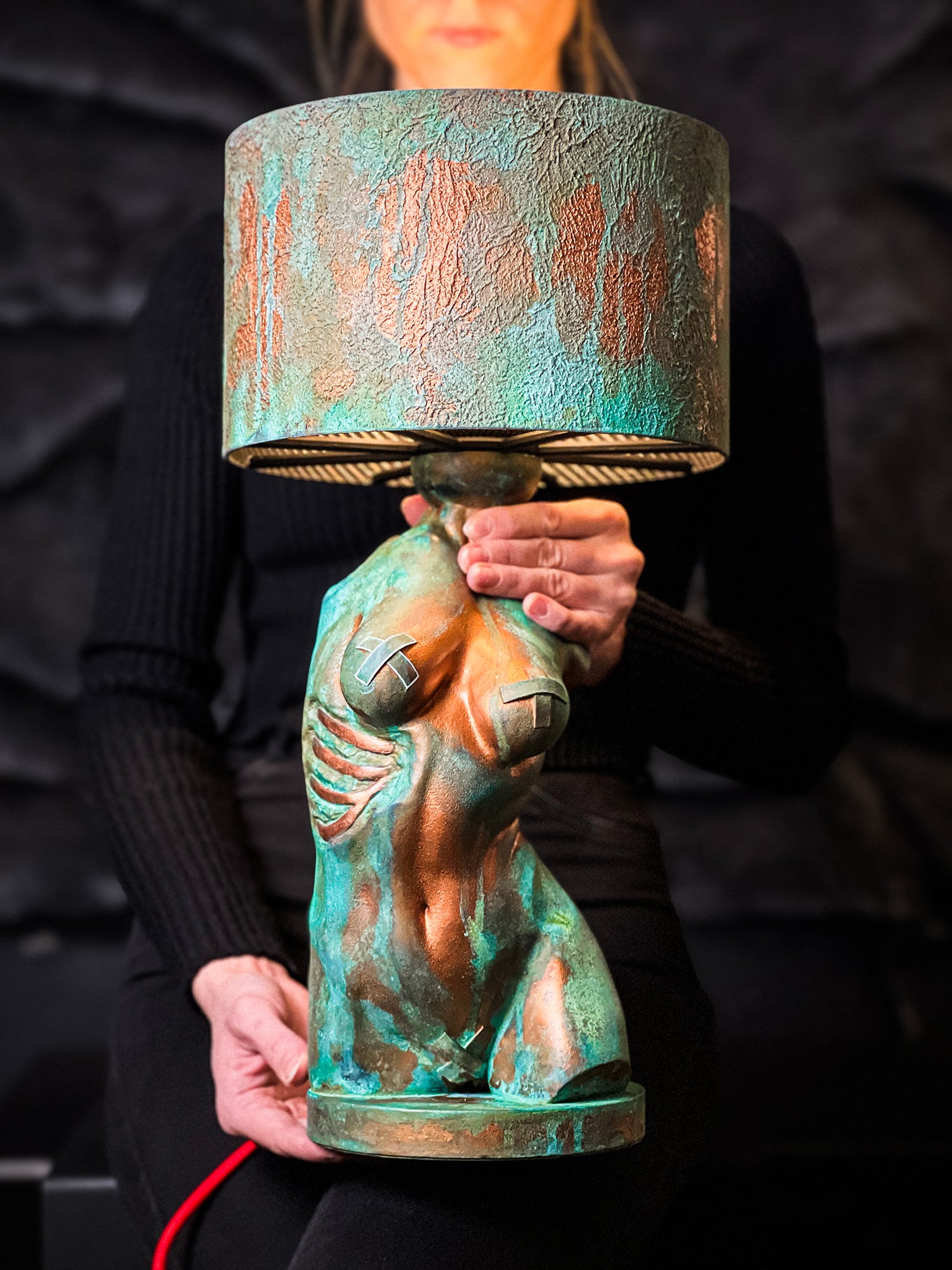 A person holds an artistic table lamp featuring a sculpted female torso base with a heavily textured verdigris and copper finish. The sculpture includes details like visible ribs and metallic tape accents over the breasts. It is topped with a matching drum shade in the same patinated style, and a bright red power cord extends from the base.