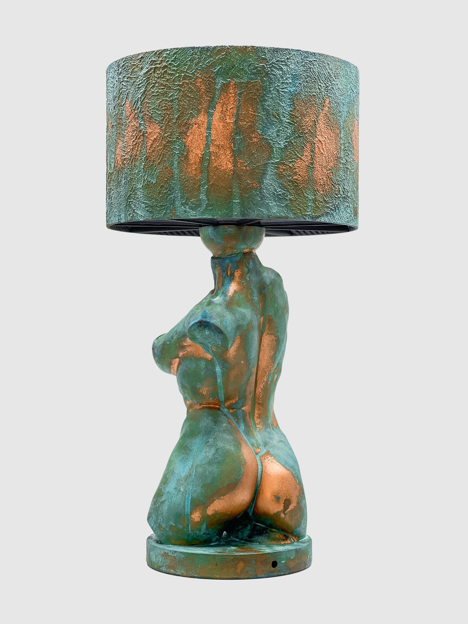 The back view of an artistic table lamp featuring a sculpted female torso base with a heavily textured turquoise verdigris and copper patina finish. The lamp is topped with a matching patinated drum shade and is isolated against a plain light gray background.