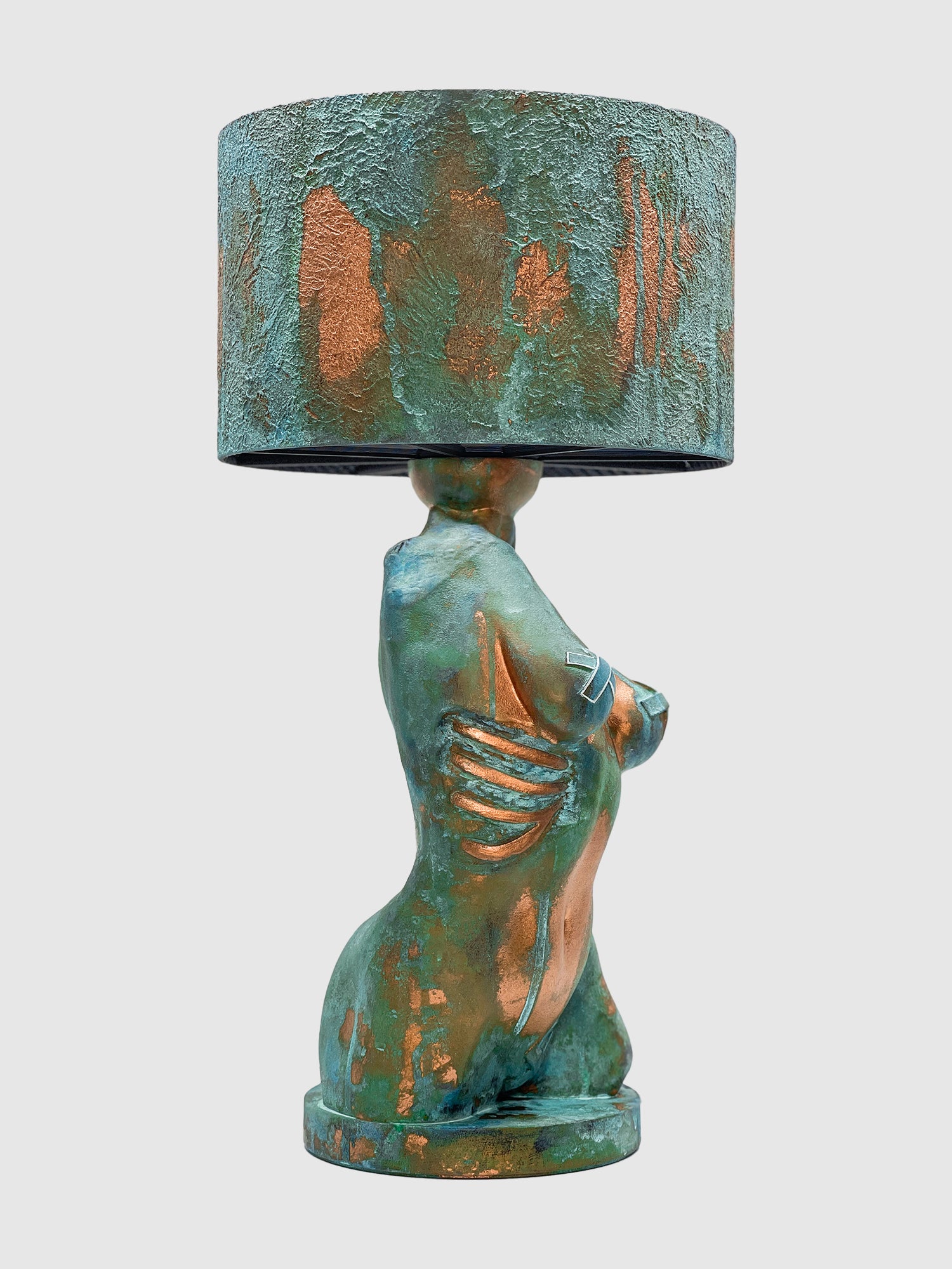 An artistic table lamp featuring a sculpted female torso base with a textured turquoise verdigris and copper patina finish. The side of the torso displays exposed copper-colored ribs, and the breasts feature metallic tape accents. The lamp is topped with a matching textured drum shade and is isolated against a plain light gray background.