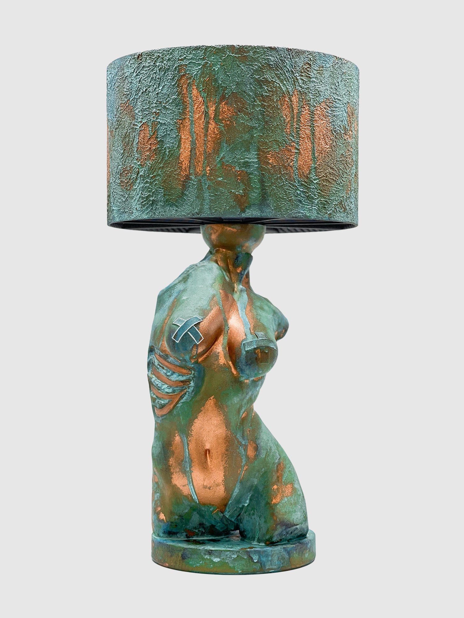 An artistic table lamp featuring a sculpted female torso base with a textured turquoise verdigris and copper patina finish. The side of the torso displays exposed copper-colored ribs, and the breasts feature metallic tape accents. The lamp is topped with a matching textured drum shade and is isolated against a plain light gray background.