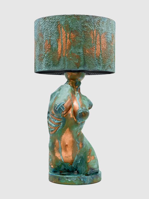 An artistic table lamp featuring a sculpted female torso base with a textured turquoise verdigris and copper patina finish. The side of the torso displays exposed copper-colored ribs, and the breasts feature metallic tape accents. The lamp is topped with a matching textured drum shade and is isolated against a plain light gray background.