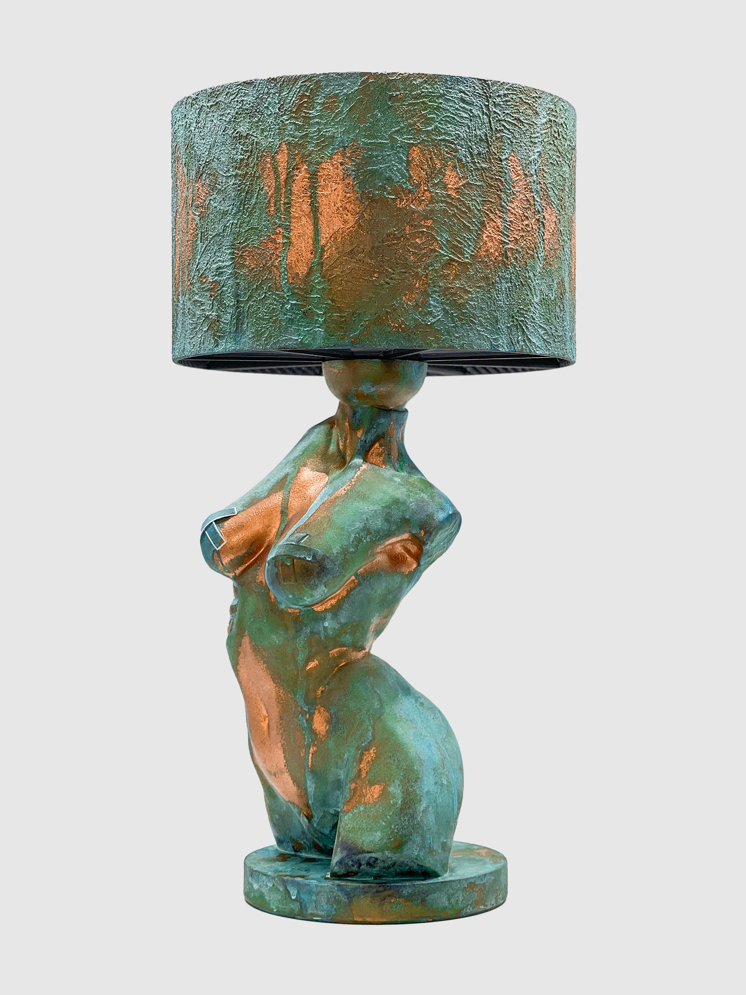 A side-angle view of an artistic table lamp featuring a sculpted female torso base with a textured turquoise verdigris and copper patina finish. The torso is depicted in a leaning pose with metallic tape accents on the breasts. The lamp is topped with a matching textured drum shade and is isolated against a plain light gray background.