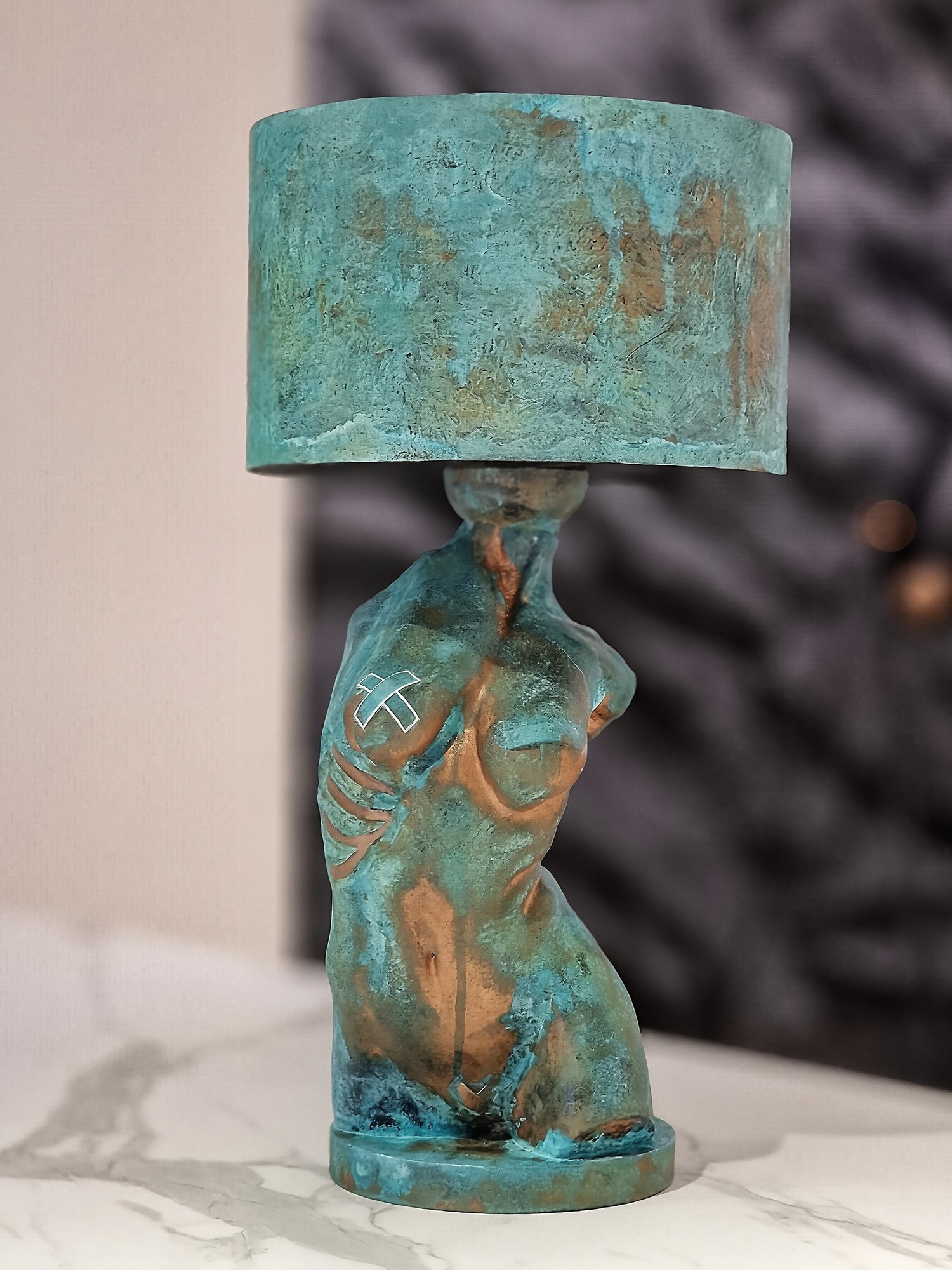 A table lamp featuring a base sculpted as a female torso and a matching cylindrical lampshade. Both the base and shade have a heavily textured verdigris patina, blending oxidized teal and turquoise with patches of metallic copper. The torso is stylized with visible ribs on one side and a small white "X" mark on the chest. The lamp rests on a white marble surface against a blurred, dark background. Created by Horror Romance.