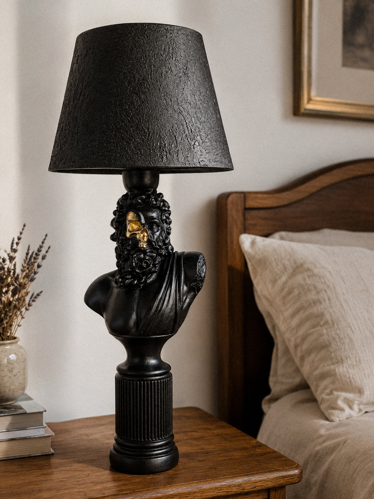 Zeus black and gold half-skull bust lamp on a wooden bedside table beside books and cream pillows.